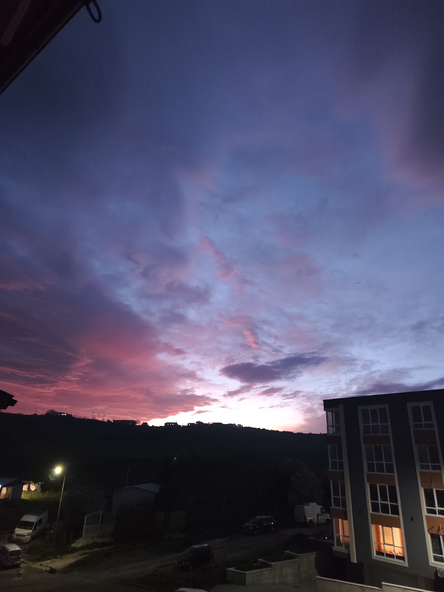 The image depicts a dusky scene, likely taken from an elevated position in Burgas, Bulgaria. The sky is dominated by a dramatic sunset with hues of purple, pink, and blue interspersed with dark clouds. A silhouetted horizon shows a line of buildings and trees on a distant hill. Closer to the foreground, there is a multi-story building on the right with illuminated windows. Below, there is a street with parked cars, a white van, and a streetlight. There is evidence of residential infrastructure with fences and possibly a building in the left foreground.