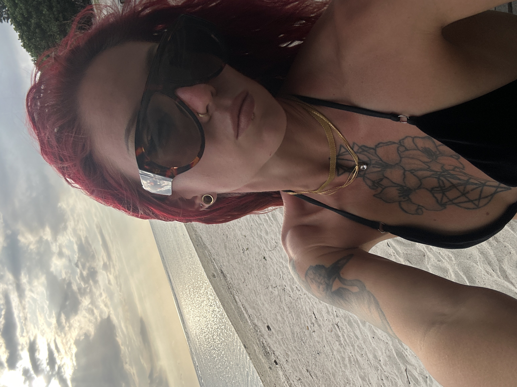 A selfie of a fair-skinned woman with red hair, wearing sunglasses, two gold necklaces and a black bikini top. She has tattoos on her chest and left arm. The background is a beach with cloudy sky and water. The location is Unknown, Indonesia.