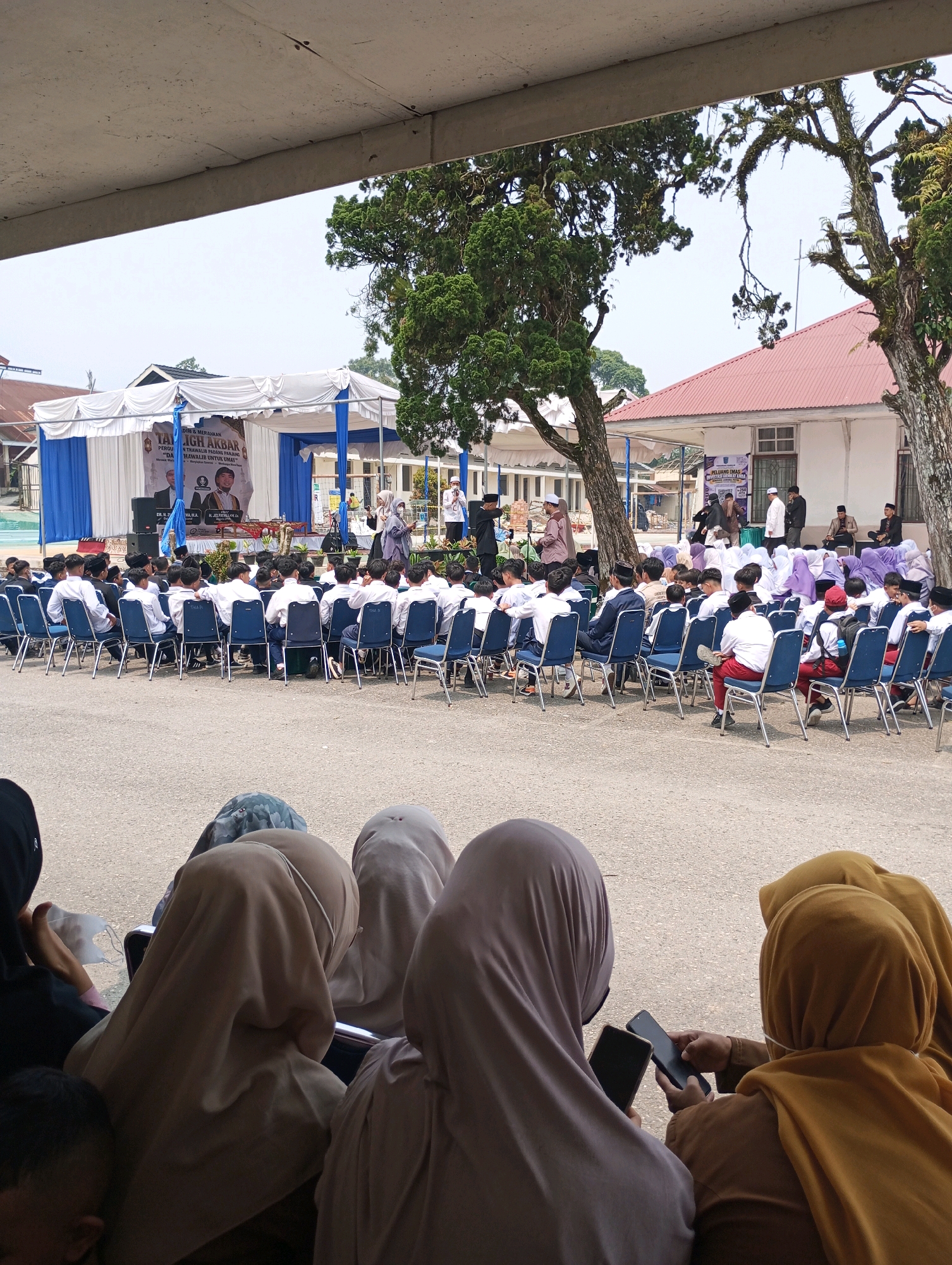 The image depicts an outdoor event taking place in the city of Padang Panjang, Indonesia. In the foreground, several individuals wearing hijabs are seated, with two of them looking at their mobile phones. Behind them, a large audience, primarily composed of young males in white shirts and dark pants or sarongs, is seated on blue chairs arranged in rows. Some are wearing caps.
In the middle ground, a stage is set up with a white and blue tent canopy. On the stage, several people are present, including individuals speaking into microphones and others listening. A banner with text in Indonesian is visible behind them, mentioning "TAJUG AKBAR" and "PERGURUAN ISLAM PADANG PANJANG" (Islamic College of Padang Panjang). There are also two portraits of men on the banner.
The background features a building with a red-tiled roof and white walls, common in Indonesian architecture. Trees with dense foliage are scattered around the area, suggesting a somewhat tropical environment. The ground is paved with asphalt. The overall scene suggests an academic or religious gathering.