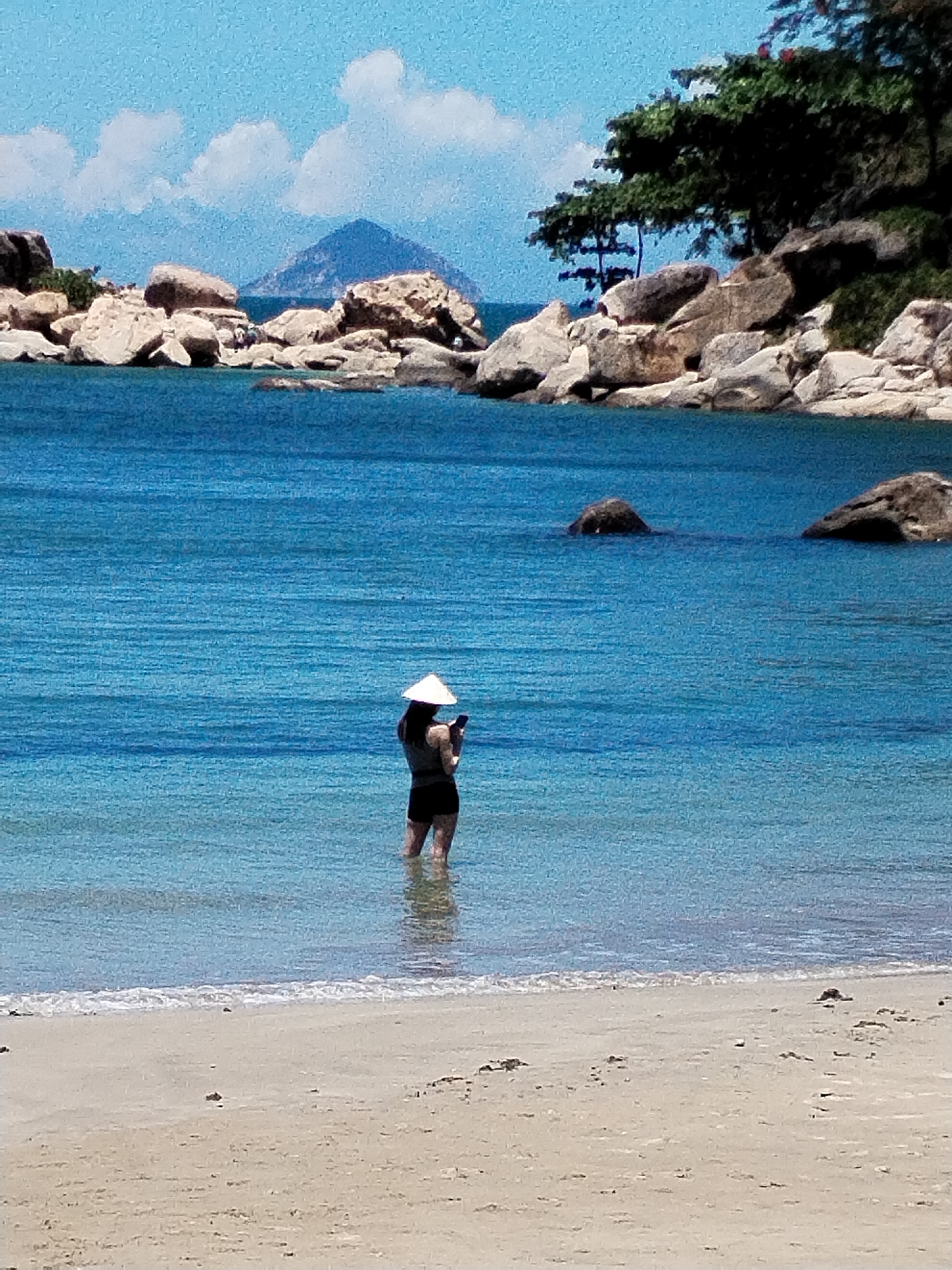 A medium shot captures a woman standing in the shallow water of a beach in Nha Trang, Vietnam. She faces away from the viewer and is wearing a traditional Vietnamese conical hat. She is using a mobile phone. The water is clear and a vibrant blue-green. The shoreline is sandy and stretches across the bottom of the frame.
In the background, rocks and trees dot the landscape, with a prominent mountain partially obscured by clouds in the distance. The sky is blue with scattered white clouds. The scene conveys a tranquil coastal environment.