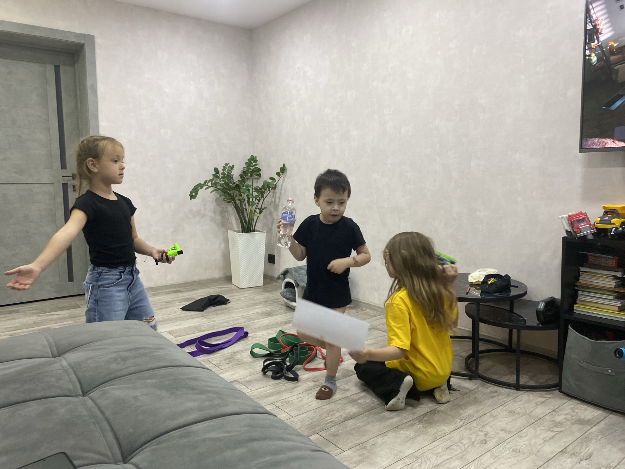 Three children are in a room with wooden floors and light-colored walls. In the foreground, a grey sectional couch is partially visible. To the left, a young girl in a black t-shirt and ripped jeans stands holding a green toy in her right hand. In the center, a young boy in a black t-shirt holds a bottle of water. To the right, a girl in a yellow t-shirt and black pants kneels on the floor. Several resistance bands in red, green, and black are scattered on the floor near the boy and girl. A purple resistance band is also visible. In the background, a doorway with a grey door is on the left, and a tall plant in a white pot is to the right of the doorway. A black table with two levels is to the right of the children, and a black shelving unit with books and a grey fabric bin is further to the right. A television is mounted on the wall above the shelving unit, and toy trucks are visible on a shelf below the television.
