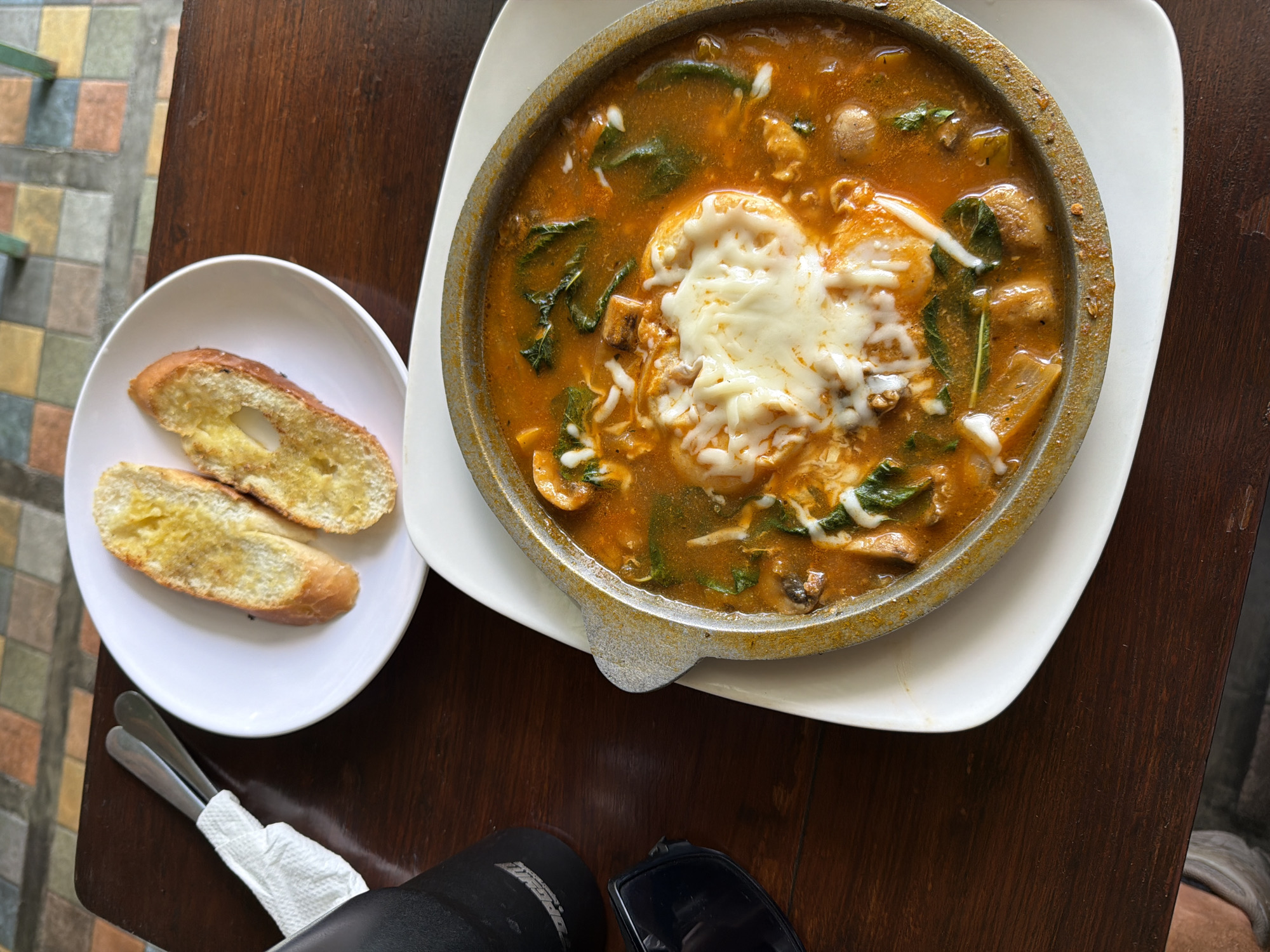 A top-down, medium shot captures a meal at an outdoor setting in North Kuta, Indonesia. A dark wood table is the backdrop. A round, cast-iron pot filled with a reddish-orange soup occupies the upper right. The soup contains mushrooms, green vegetables, and is topped with melted white cheese. To the left, a white plate holds two slices of bread. A napkin-wrapped set of silverware rests near the plate. In the lower part of the frame, a black object, along with sunglasses, are visible on the table. The foreground features a patterned floor of colored tiles.
