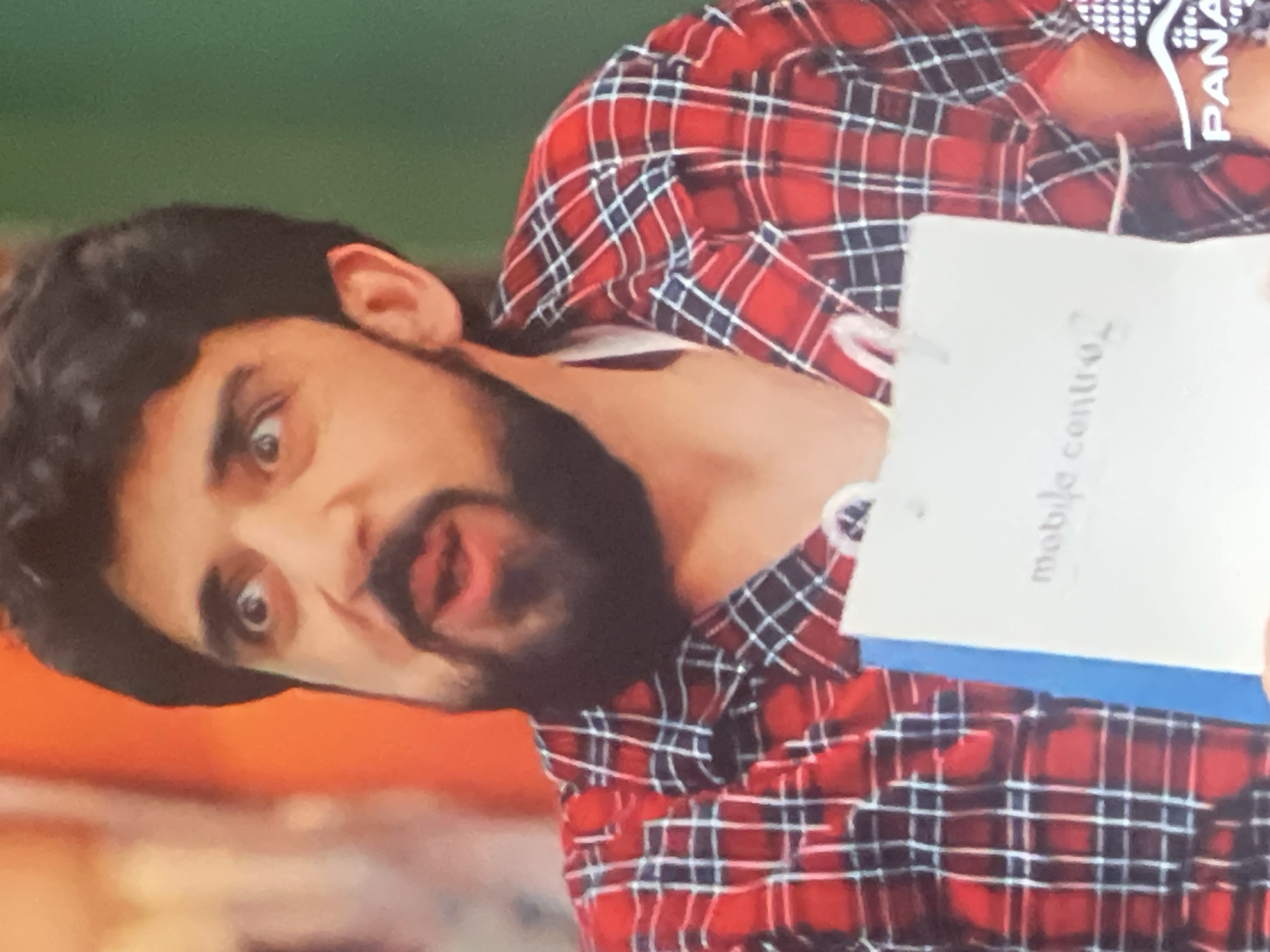The image shows a close-up of a man with a beard and dark hair, wearing a red and black plaid shirt. He is holding a white paper bag with text that appears to say "mobile centro". The man has a surprised or shocked expression on his face, with his eyes wide open and his mouth agape. The background is blurred, with patches of orange and green. There is also a logo in the bottom right corner with the word "PANA". The scene does not contain any specific landmarks or details that would allow for identification of a location in Vanadzor, Armenia.