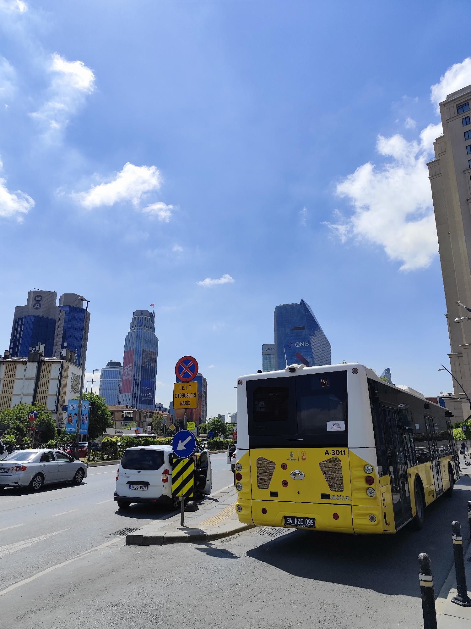 The image captures a street scene in an urban environment. In the foreground, there is a yellow and white bus parked on the right side of the road, its license plate reads "34 HZC 099" and A-3011 is written on the bus. A white van with an open door is parked near a street sign and another car is also visible. The street sign includes a no-entry symbol with the inscription "I.E.T.T. OTOBUSLERI HARIC". In the background are several modern skyscrapers, including one with the "QNB" logo. The sky is bright blue with scattered white clouds.