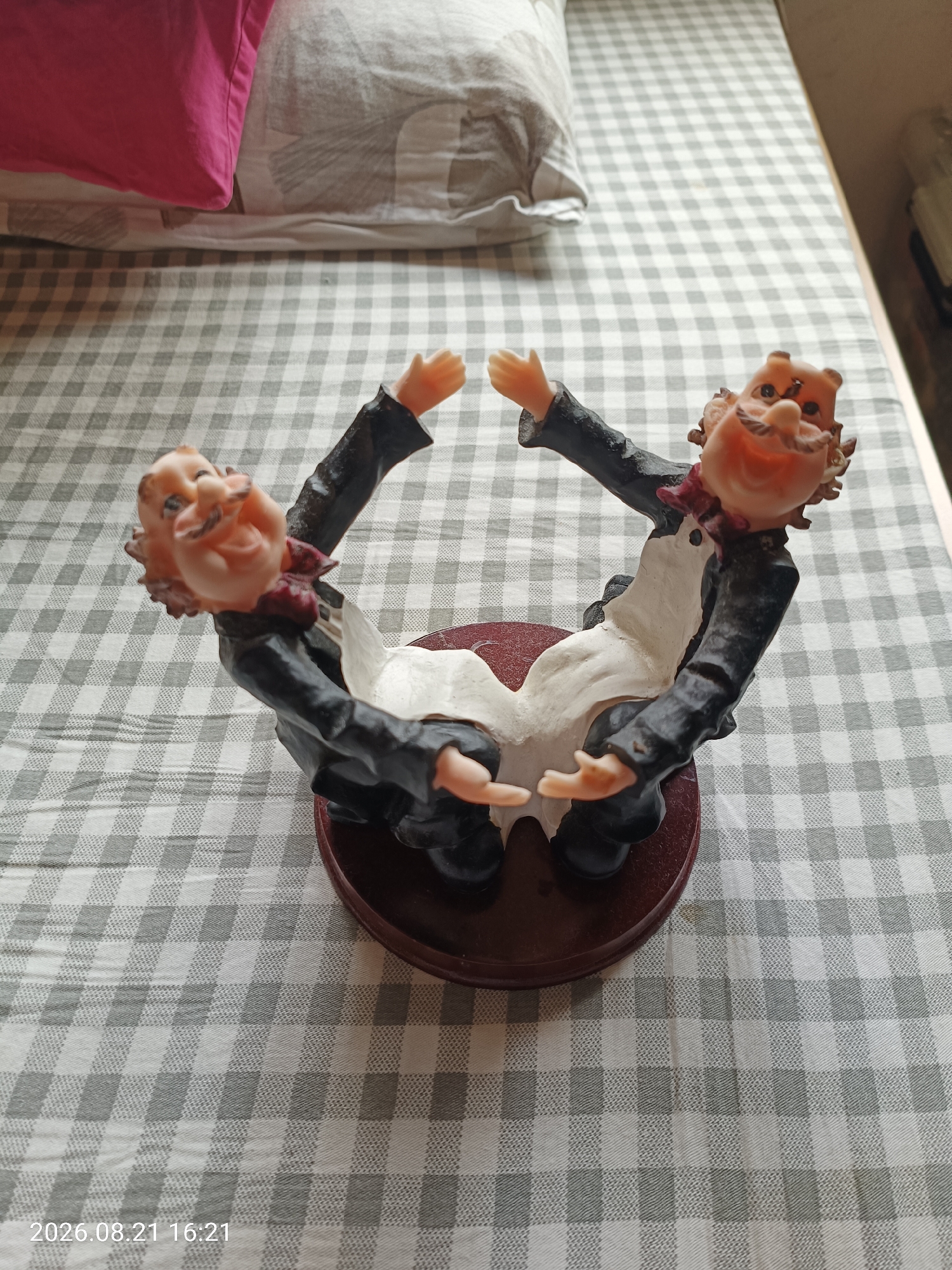 The image shows a novelty figurine of two cartoonish men with exaggerated features, possibly wine bottle holders. They are depicted with large heads, mustaches, and are dressed in formal attire with white shirts and black jackets. Their arms are extended outwards, forming a frame. The figurine is placed on a dark red, circular base. The background consists of a checkered tablecloth with a pattern of light and dark grey squares. A pink pillow is visible in the upper left corner of the image. The timestamp "2026.08.21 16:21" is present in the lower left corner. The image does not contain any recognizable landmarks or features that would indicate its location in Hong Kong, China.
