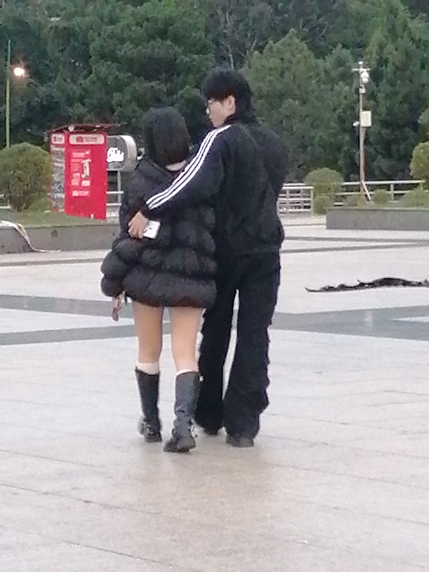 In Da Lat, Vietnam, a couple walks away from the camera on a paved outdoor area. The person on the left, likely female, wears a black puffer jacket, white socks, and black boots. They are holding a white smartphone in their right hand. The person on the right, likely male, wears a black tracksuit with white stripes on the sleeves and black pants. The background features lush green trees and a bright red kiosk with signage. The ground is a light-colored stone or concrete pavement with visible lines.