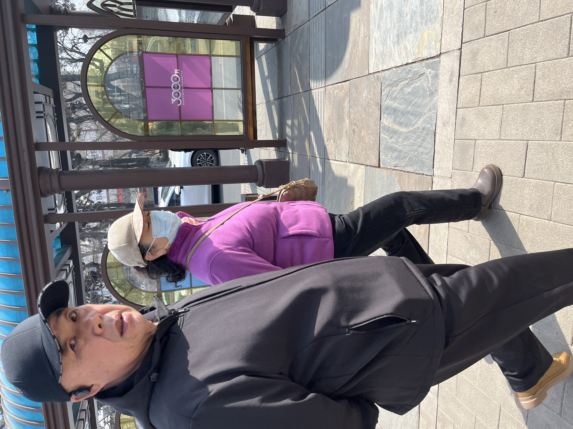 In Changchun City, China, a man and woman are walking along a paved walkway near a bus stop. The man is wearing a black baseball cap and a black jacket. The woman is wearing a pink jacket and a beige hat with a white face mask. The bus stop has a sign with the number "3000" visible. The walkway is made of concrete slabs and brick.