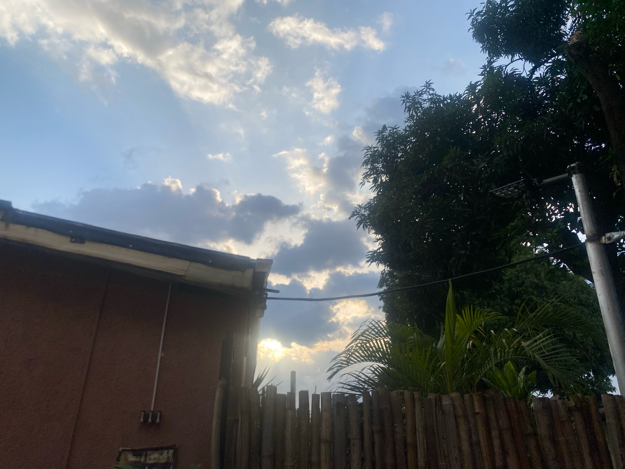 In Barden Yana, Nigeria, an outdoor scene captures a building with a brown, textured wall and a white gutter along its roofline. In the foreground, a fence constructed from vertical bamboo poles separates the property. Beyond the fence, lush green foliage, including palm fronds and the dense canopy of a large tree, is visible against a sky filled with scattered clouds and rays of sunlight breaking through. A portion of a utility pole with some equipment is also present on the right side of the frame. The overall impression is a view from a residential or semi-rural area, possibly late afternoon due to the quality of light.