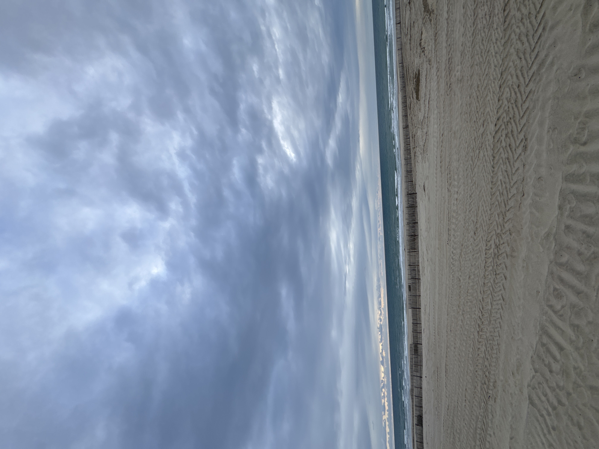 The image depicts a beach scene in Soulac-sur-Mer, France. The sky is predominantly filled with heavy, layered clouds. A thin strip of ocean is visible between the clouds and the sandy beach. The beach is marked with tire tracks. A fence borders the beach. No people are visible in the image.
