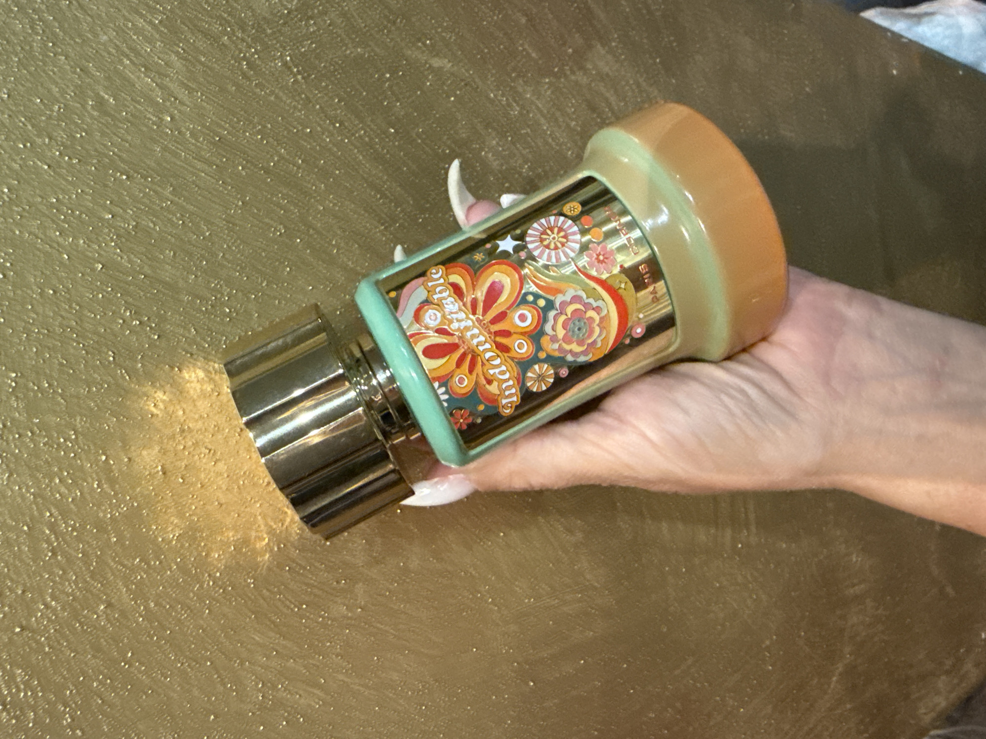 A hand holds a bottle of perfume against a gold, textured background. The perfume bottle is cylindrical with a metallic gold cap and a light green glass body decorated with colorful floral and paisley patterns in shades of orange, red, yellow, and pink. The word "indomitable" is visible in a stylized font on the bottle. The hand has manicured nails with white polish. The background is a mottled gold surface with a speckled texture. There is no discernible location in the city of Odesa, Ukraine.