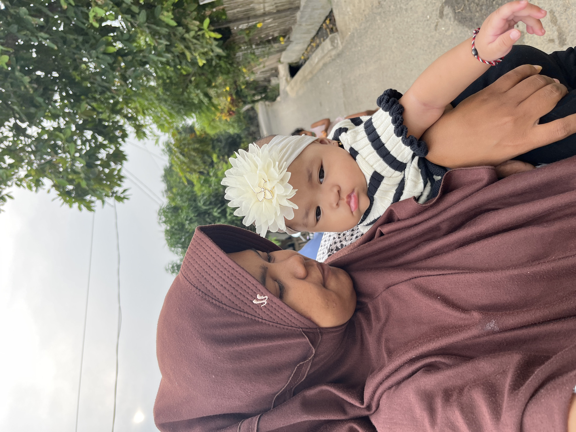 In Kruengmate, Indonesia, a woman wearing a maroon hijab holds a baby. The woman, in the foreground, looks down at the baby with a thoughtful expression. The baby, adorned with a white flower headband and a black and white striped shirt, gazes forward with a pout. The background shows a street lined with trees and a fence, suggesting a residential area.