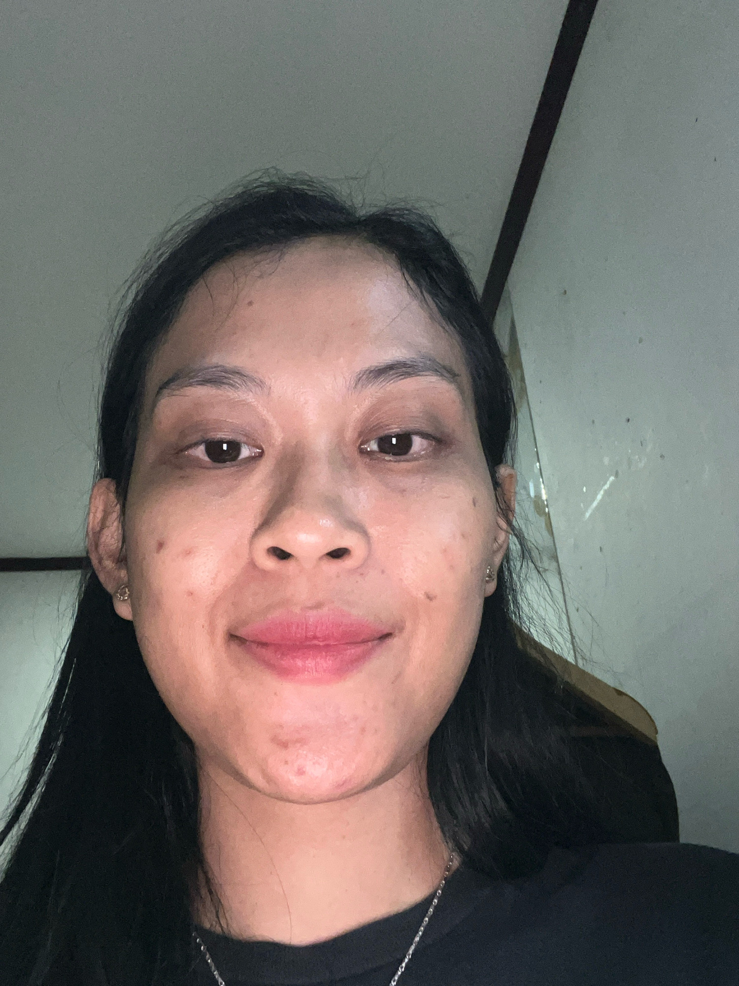 A close-up, selfie-style, head-and-shoulders portrait of a young adult Indonesian woman is captured in a dimly lit indoor setting. The woman has long, dark hair that frames her face and extends down her shoulders. She is looking directly at the camera with a gentle smile. Her complexion is visible, with some natural skin texture and subtle makeup. She is wearing a dark-colored shirt, and a silver chain necklace is visible around her neck. The background appears to be a wall with a dark vertical element, possibly a doorframe or corner, and a lighter, textured surface. The lighting is soft, coming from above and slightly to the side, casting gentle shadows. The overall impression is of a casual, personal moment. The scene appears to be an interior residential or communal space within Kecamatan Pati, Indonesia.