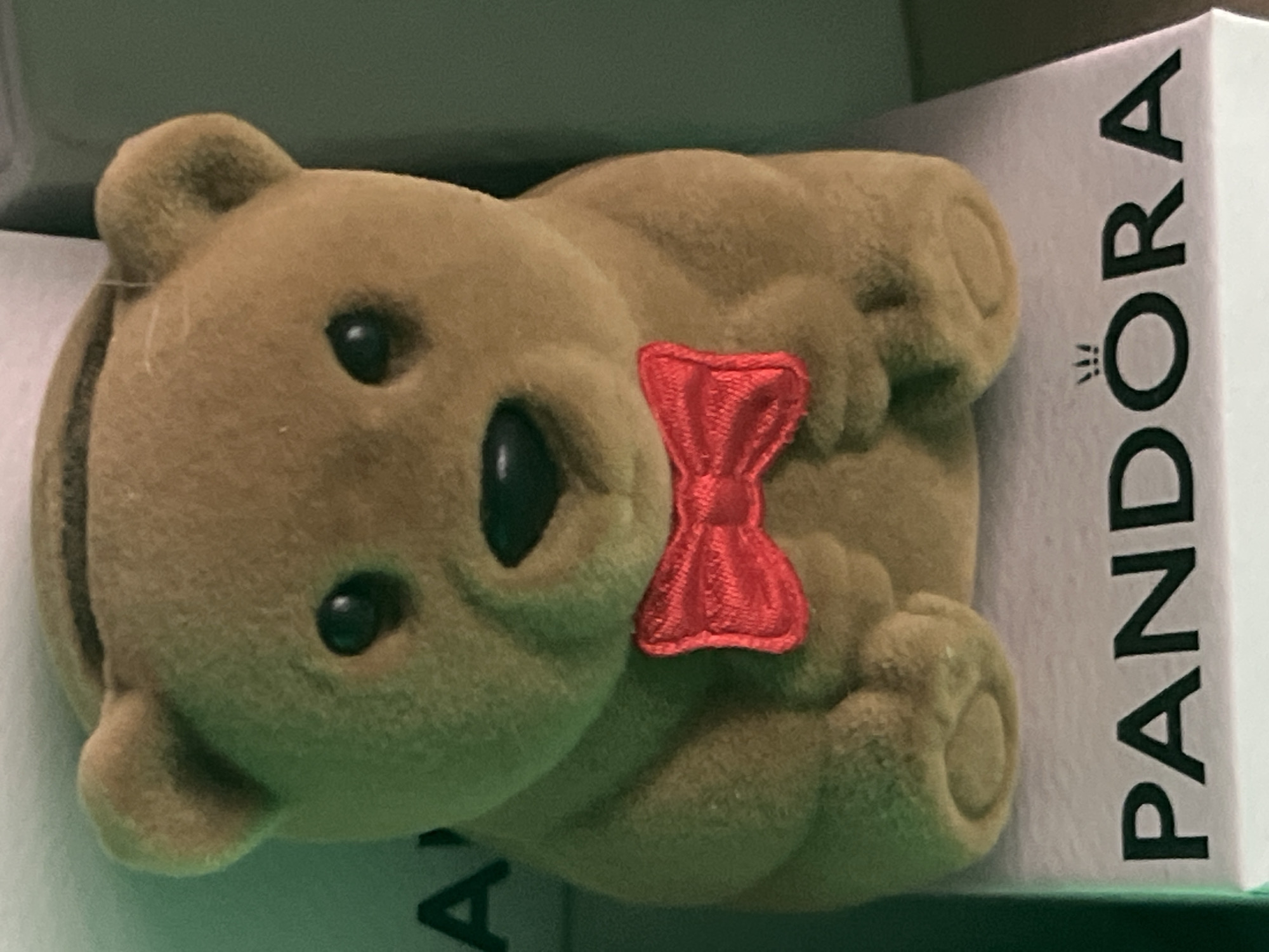 A close-up, eye-level shot captures a plush, tan teddy bear wearing a red bow tie. The bear is positioned on top of a white box with "PANDORA" printed in black lettering across its surface. The background is dark, with a green hue on the left side and a darker shade on the right, suggesting an indoor, possibly dimly lit environment. The bear's eyes are black and beady, and its nose is a small, black dot. Its ears are rounded, and its paws are resting on its lap. The overall impression is that of a decorative item or a collectible.