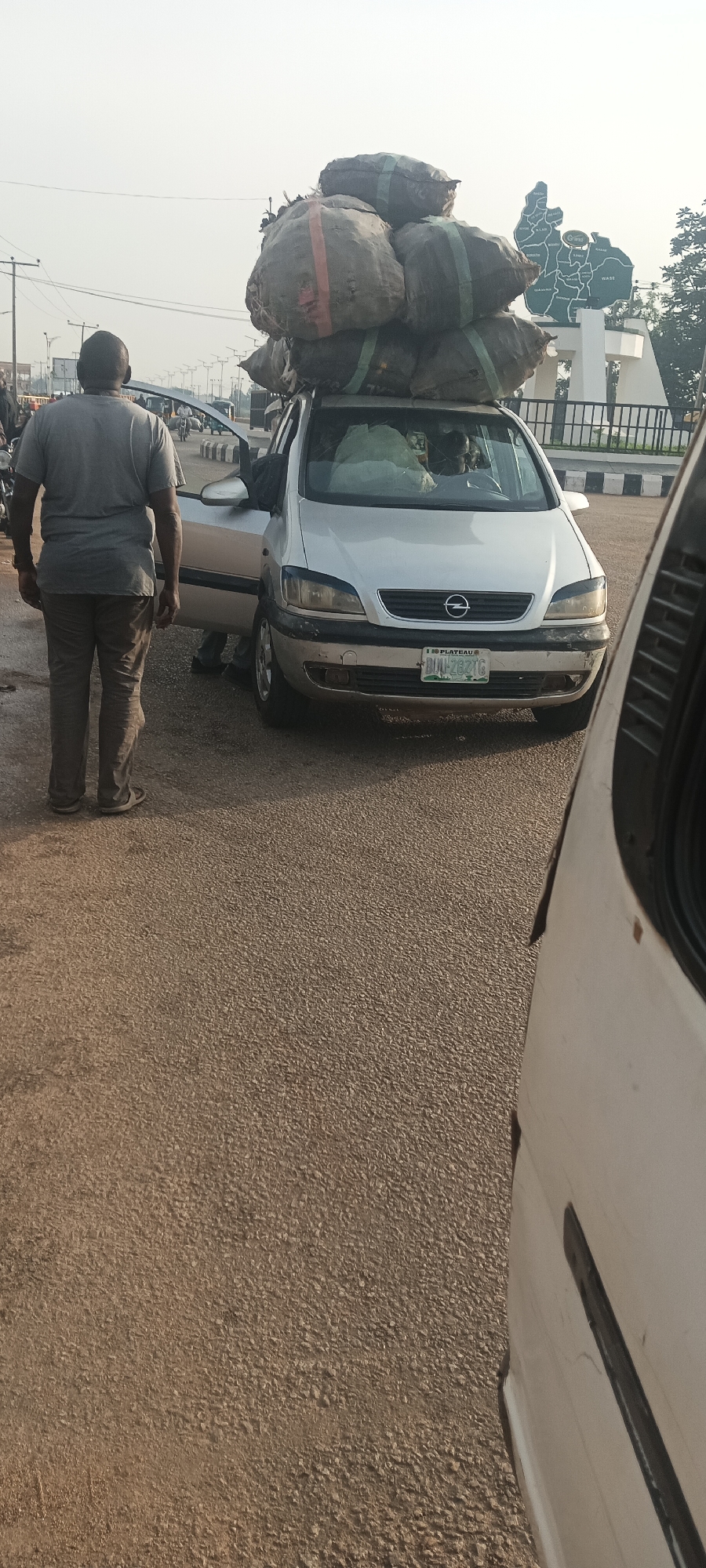 A silver Opel Zafira car is stopped on a road in Bukuru, Nigeria. The car is heavily laden with at least six large sacks secured on the roof. A man in a grey shirt and brown trousers stands near the open driver's side door of the vehicle. The car has a "PLATEAU" license plate with the number "BUU-282 TG". In the background is a monument resembling a map of a region, along with road barriers. Part of another white vehicle is visible on the right side of the frame. The road surface is asphalt.