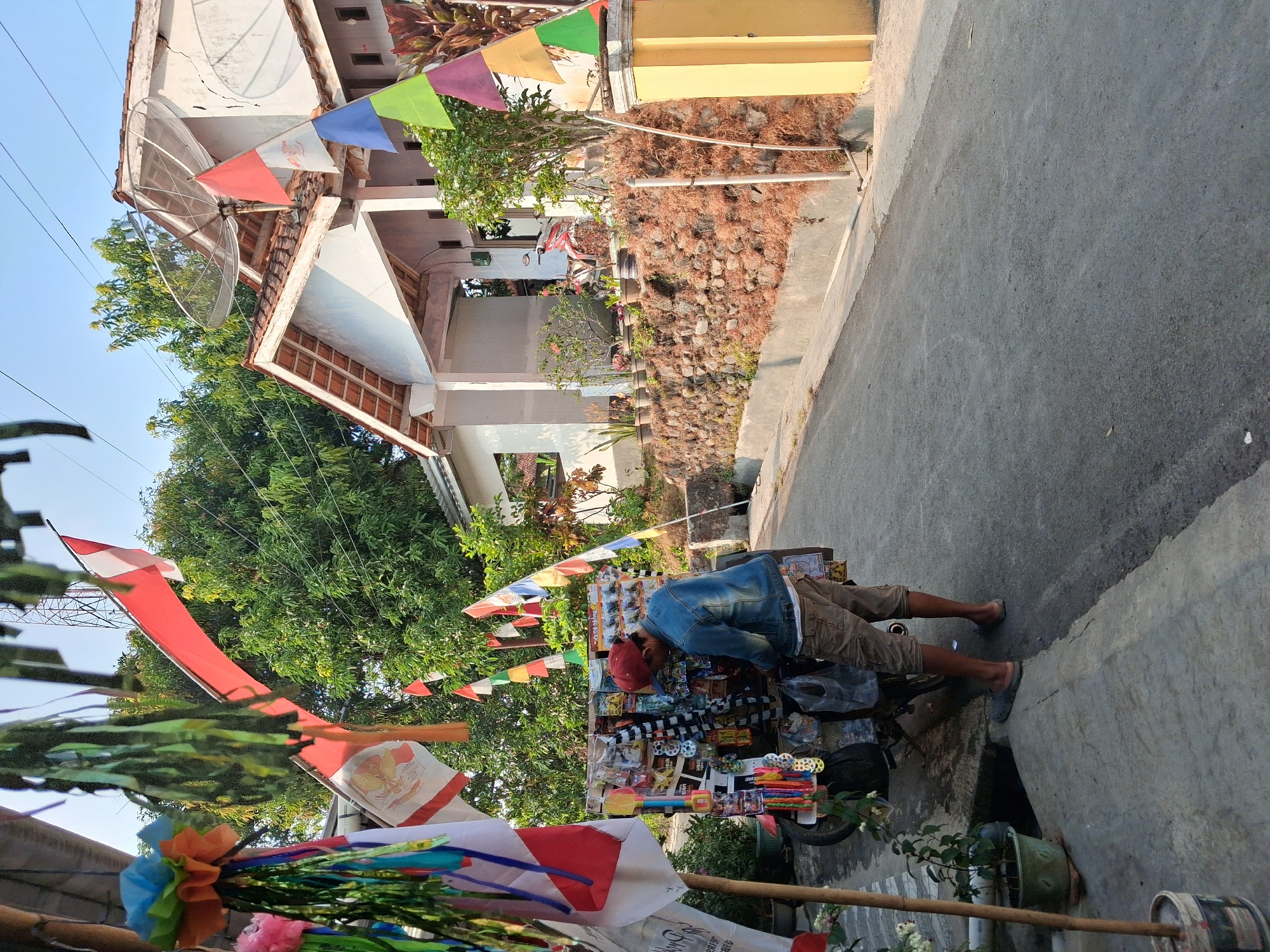 This image depicts a street scene in Bantarkawung, Indonesia, likely during a national celebration given the prevalence of Indonesian flags. In the foreground, a man wearing a denim jacket, beige cargo shorts, a red baseball cap, and sandals is browsing a small mobile stall. The stall, mounted on a motorcycle, displays various items, including what appear to be toys and accessories. Behind the stall, several Indonesian flags and decorative banners are visible, including the national red and white flag and smaller pennants in red, white, blue, green, and yellow. The stall is situated on a paved street, with a concrete walkway on the left and a dry stone wall bordering the right side of the street. A white house with a satellite dish on its roof is visible in the background, with trees and potted plants surrounding the property. The sky is clear and blue, suggesting it is daytime. The scene conveys a sense of local commerce and festive atmosphere within a residential area.
