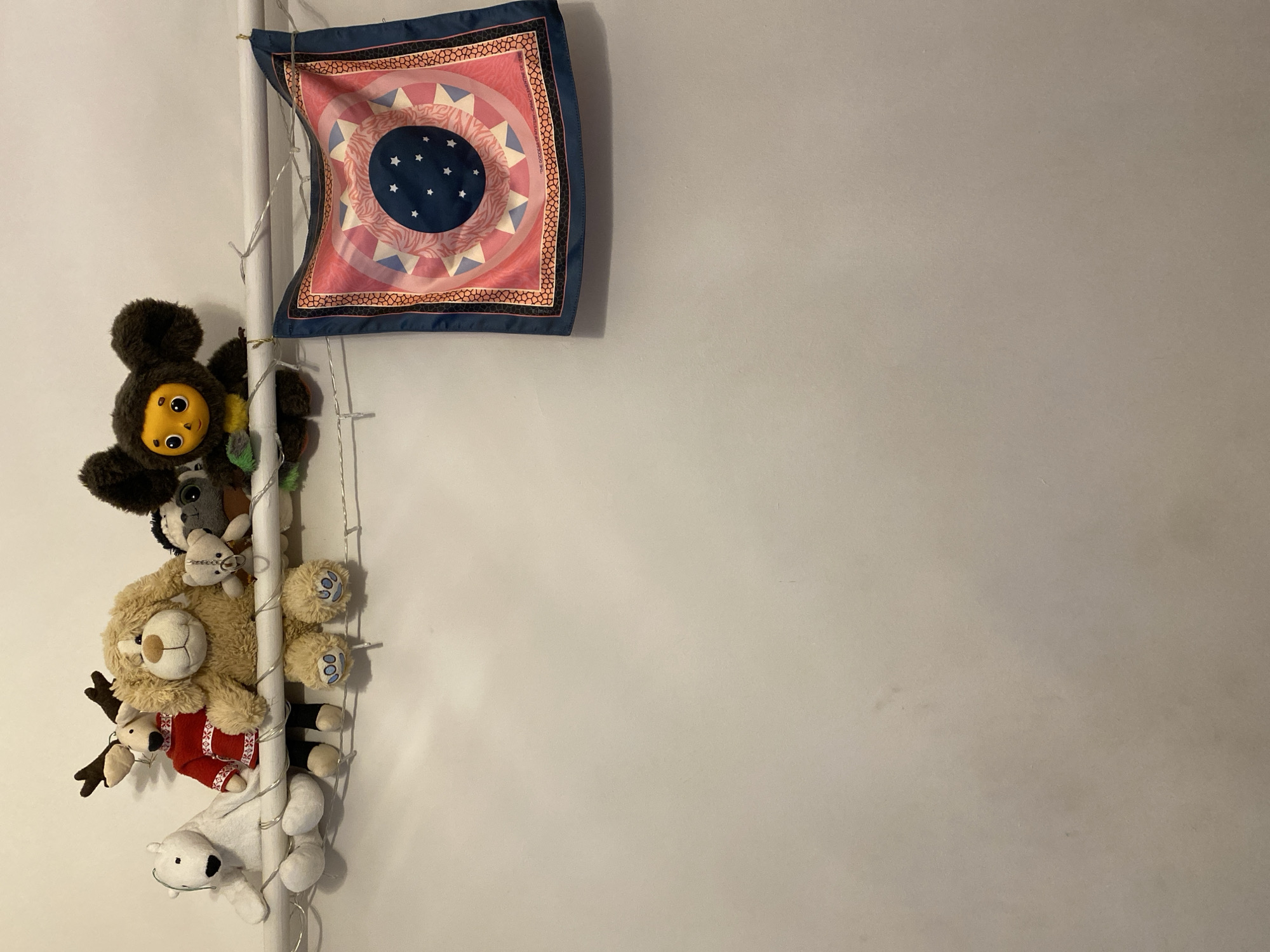 In the city of Beyoğlu, Turkey, a white shelf is mounted on a textured white wall. The shelf holds several plush toys, including a white polar bear, a reindeer in a red sweater, a fluffy brown dog, a smaller brown bear, and a dark-colored monkey-like character. A string of small, fairy lights is draped along the shelf and the wall below it. To the right of the toys, a colorful scarf with a circular pattern is hanging from the shelf. The scarf is predominantly pink and blue with star-like shapes and geometric designs.