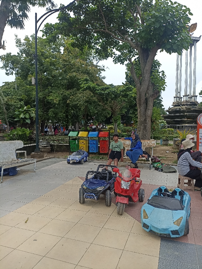 This image captures a vibrant outdoor public park scene, likely in Tulungagung, Indonesia, as suggested by the context and visual cues. The setting features paved pathways, lush greenery, and various recreational elements.
In the foreground, a paved area displays several electric ride-on toy vehicles: a blue off-road buggy, a red motorcycle, and a light blue sports car. Another blue toy police car is visible further back. These toys suggest a recreational or rental activity for children.
Several people are present in the park. In the mid-ground, a young boy in a green t-shirt and black shorts sits on a concrete bench alongside a woman dressed in a blue hijab and blue clothing. On the far right, other individuals, including one wearing a hat, are seated on small stools. More people can be seen in the background, scattered among the trees and park features.
A notable feature in the mid-ground is a set of four colorful trash bins (yellow, red, blue, green), clearly labeled "DAUR ULANG" (Recycle) with distinct icons for different waste categories (organic, plastic, paper, metal). Behind these bins, a water feature or pond with lily pads is visible.
The park is abundant with large, leafy green trees, providing extensive shade. A tall black light pole stands on the left side of the frame. In the background on the right, a prominent monument or sculpture with several tall, white pillars and a bird-like figure on top (possibly a Garuda, a national symbol in Indonesia) stands as a focal point.
The scene takes place during daytime, under what appears to be an overcast or partly cloudy sky, providing soft, diffused light. The overall mood suggests a relaxed and leisurely atmosphere in a public recreational area. Partial text "ENSUP" is visible on a sign near the bench on the left, while small, unreadable text appears on a red and white sign on the far right.