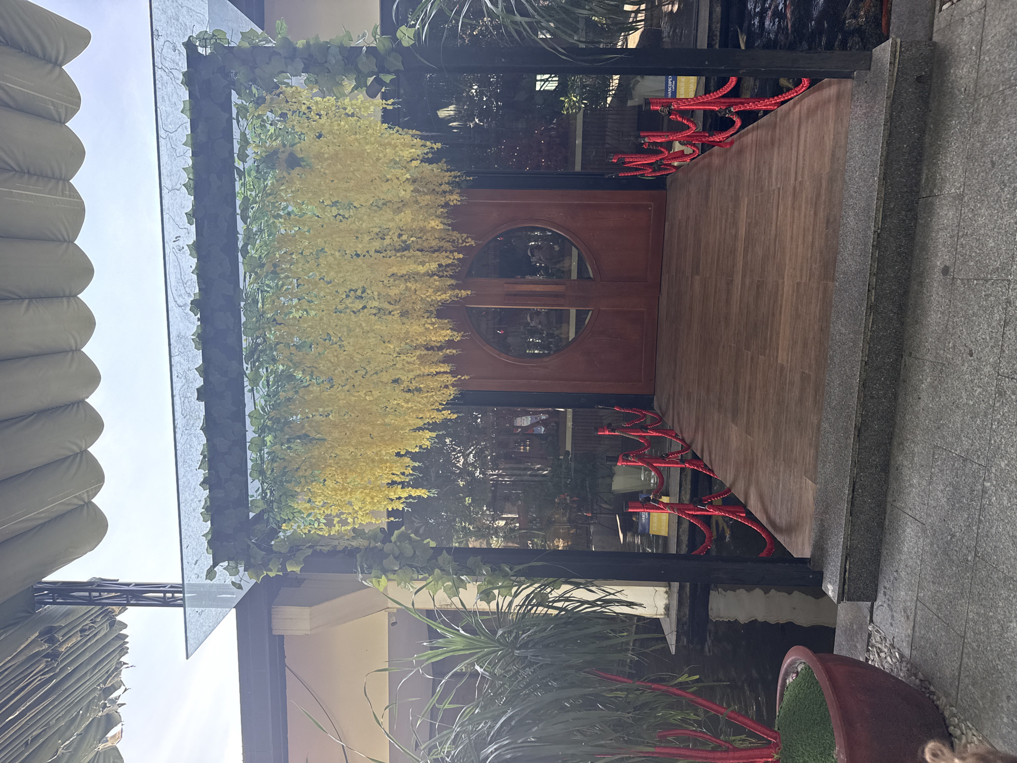 The image shows an outdoor entrance to a building, decorated with hanging yellow flowers and ivy. A wooden walkway leads to a pair of wooden doors with a circular glass insert. Red ropes on stanchions line the sides of the walkway, bordering a dark body of water that appears to contain koi fish. Large green plants are positioned to the left of the entrance. The architectural style suggests a restaurant or a similar establishment. The overall scene is well-maintained and visually appealing, likely designed to create an inviting atmosphere for visitors. The location is identified as Nam Nha Trang Ward, Vietnam.
