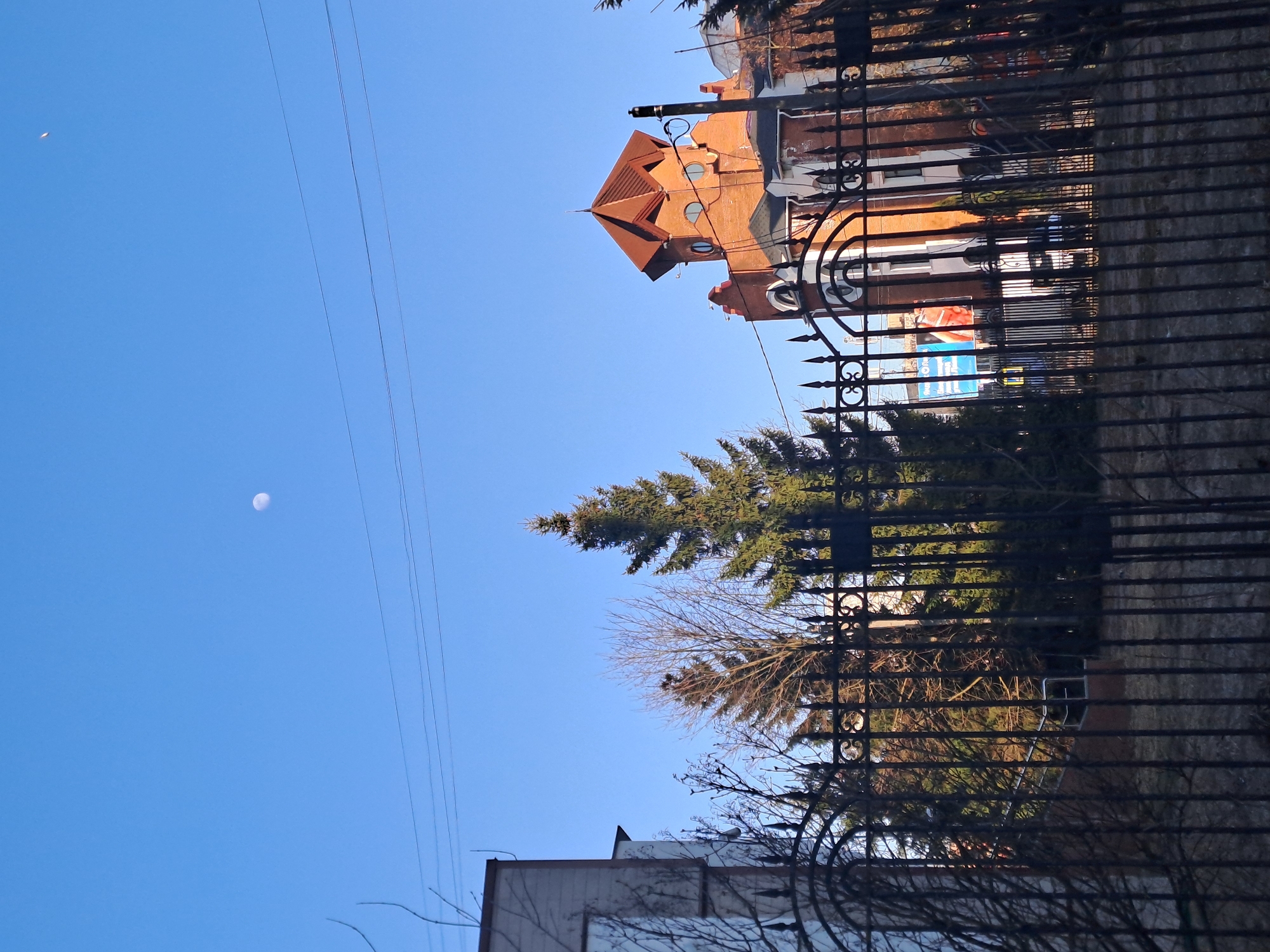 The image depicts an upward-looking view from behind a black metal fence in Elektrostal, Russia. In the top portion of the image, a red brick building with a distinctive turret and round windows is visible. Below the building and partially obscured by the fence and trees, a billboard is visible. A tall evergreen tree stands between the fence and the building. In the upper-left of the image is a blue sky with a visible crescent moon. Power lines stretch across the sky. In the lower portion, a white building and bare tree branches are visible.