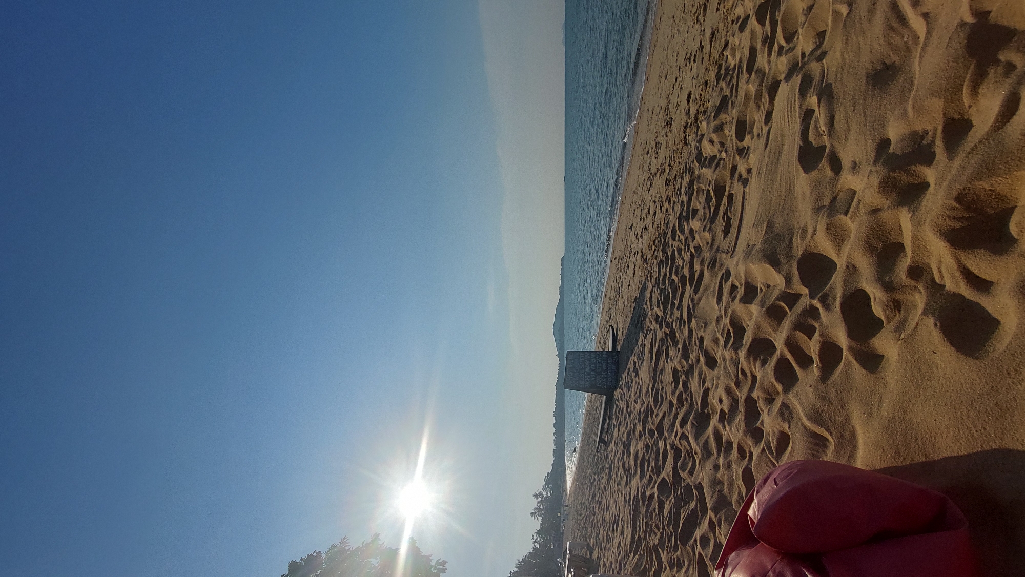 This image captures a bright, sunny beach scene, likely in Baan Mae Nam, Thailand, presented with a vertical orientation. The clear blue sky occupies the entire left side, with a brilliant sunburst emanating from the lower-left corner, casting strong rays across the frame.
To the right, the calm blue water of the ocean meets the golden-brown sand of the beach, which dominates the right half and foreground of the image. The sand is rich in texture, covered with numerous footprints and undulations, creating a pattern of light and shadow indicative of a low sun angle. Along the distant horizon line, a faint landmass or coastline is visible across the water.
On the beach, near the water's edge and slightly right of center, stands a dark, A-frame sign, resembling a chalkboard. While the white text on its surface is not fully legible, it appears to list items or information, suggesting a beachside establishment. In the far bottom right corner, the vibrant red fabric of two soft beanbag chairs or similar seating is visible, hinting at a leisurely and comfortable setting.
The scene suggests a peaceful moment during the golden hour, either early morning or late afternoon, characterized by clear weather and abundant sunshine. The overall mood is serene and inviting.