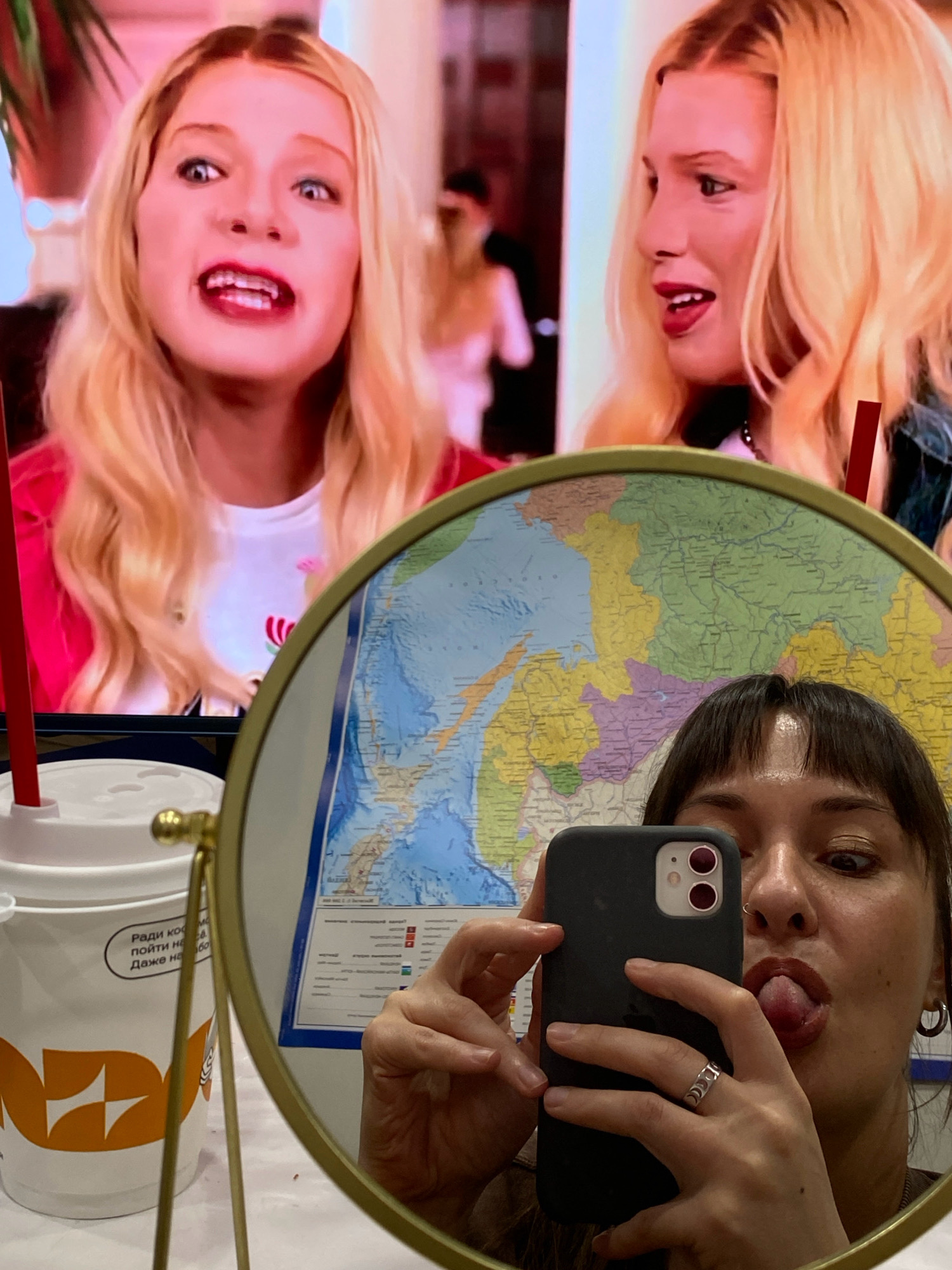 The image shows a woman taking a selfie in a mirror. She is sticking her tongue out and holding a smartphone. In the background, a map is visible on the wall. There are also a cup with a straw and a stand for the mirror. The scene appears to be indoors, and there is no indication of the city of Votkinsk, Russia, being present in the image.