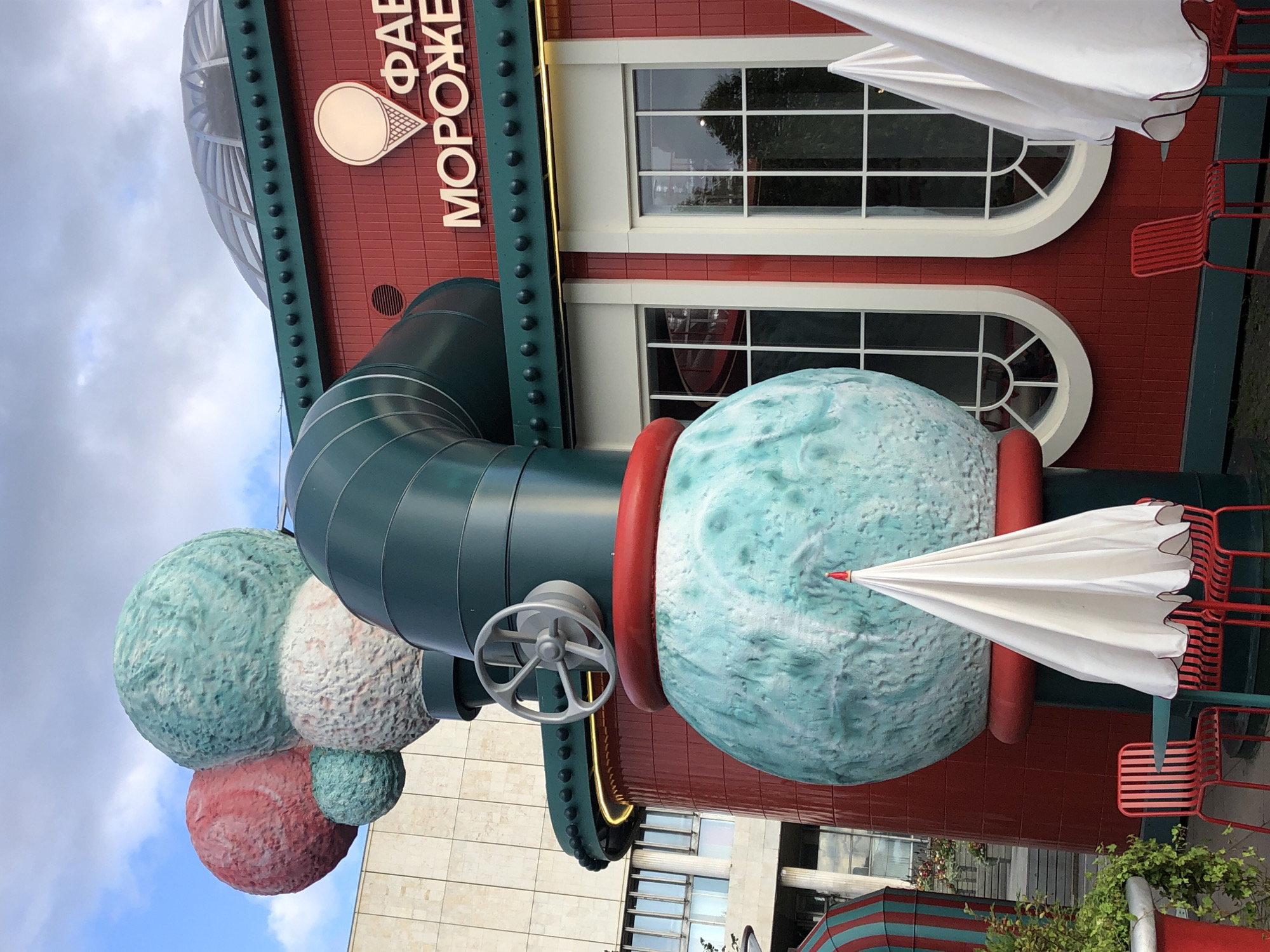 The image shows the exterior of an ice cream factory building in Moscow, Russia. The building is red with white trim and features large windows. A prominent sculpture of ice cream scoops and pipes is attached to the side of the building. Several white umbrellas and red chairs are set up outside the building, suggesting an outdoor seating area. The sky is partly cloudy. The signage on the building appears to be in Russian and includes the word "Мороженое," which means ice cream.