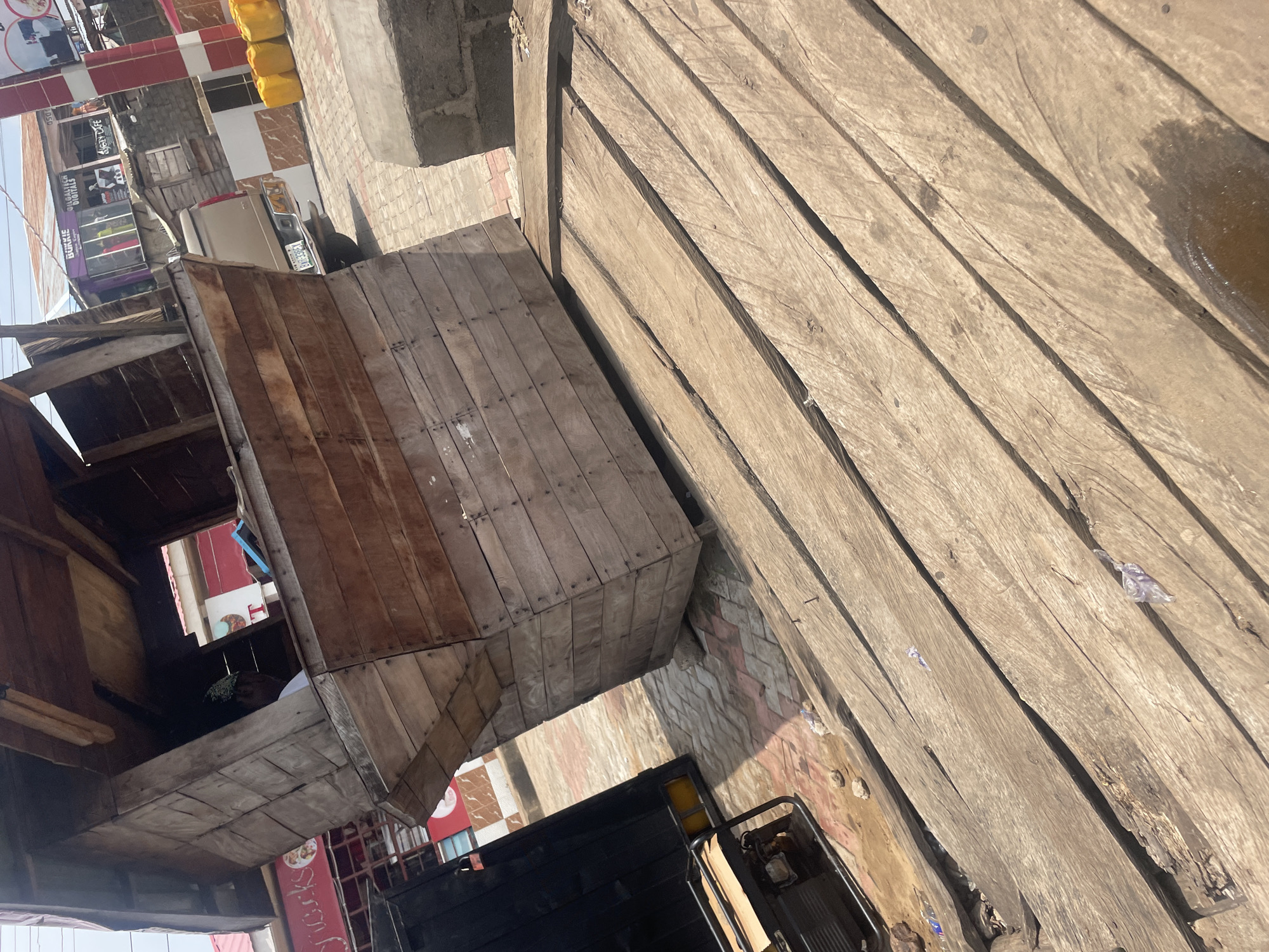 The image depicts a street scene in Lokoja, Nigeria, characterized by a weathered wooden stall tilted on a wooden platform. The stall has a rustic appearance, constructed from planks of varying shades of brown. In the background, a beige pickup truck with a license plate is visible, alongside several yellow containers and a building with red and white striped pillars. Adjacent to the stall, a black object, possibly a generator or a vehicle, is partially visible on the left. The ground is paved with interlocking bricks, and there is debris scattered around. The lighting suggests it is daytime, with shadows indicating the sun's position. The overall impression is of a typical urban setting in Lokoja, possibly a marketplace or a roadside vendor area, with signs of commercial activity and everyday life.