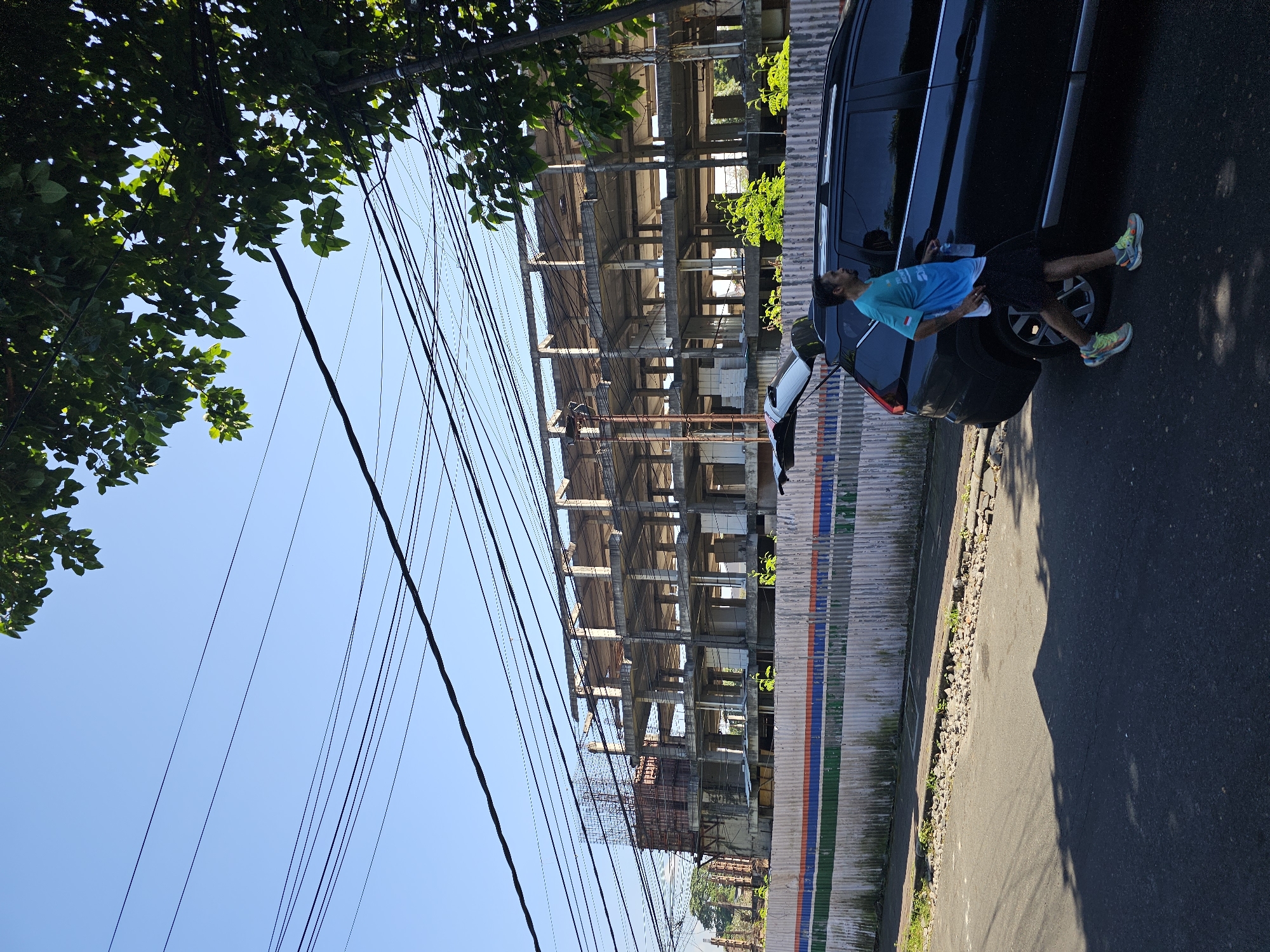 A man in a teal and white t-shirt and black shorts walks on a paved street next to a dark SUV with its trunk open. The street is lined with a corrugated metal fence with horizontal stripes of blue, white, orange, and green. Behind the fence, a multi-story concrete building is under construction. The sky is clear and blue. Overhead, a dense network of electrical wires crisscrosses the scene, partially obscured by the green leaves of a tree in the upper right corner. The image appears to have been taken in Denpasar, Indonesia, based on the context of the surroundings.