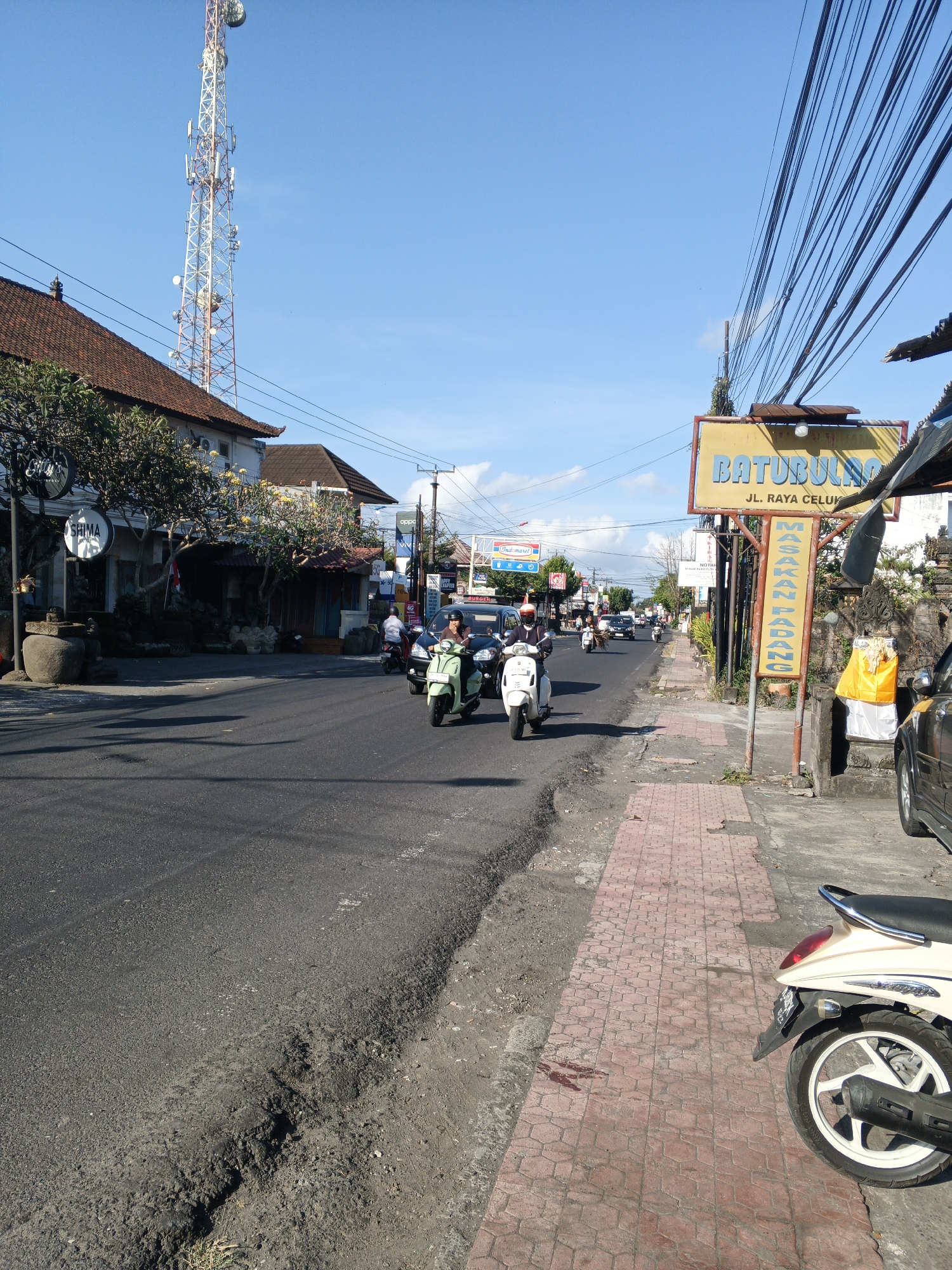 The image depicts a street scene in Sukawati, Indonesia. The road is paved with asphalt and shows signs of wear and tear, with a noticeable pothole on the right side of the foreground. A sidewalk with red patterned paving stones runs alongside the road.
On the sidewalk, there are several objects. A yellow sign with red lettering that reads "BATUBULAN JL. RAYA CELUK" is prominent. Below this, a vertical yellow sign with black lettering states "MASAKAN PADANG". Adjacent to these signs, a decorative stone structure is draped with a yellow cloth. Further down the sidewalk, a light-colored scooter is parked.
The street is active with vehicular traffic, primarily scooters. A person is riding a light green scooter in the foreground, while other scooters are visible further down the road. A black car is parked on the right side of the street.
Buildings line the left side of the street. One building has a traditional tiled roof and features trees with yellow flowers in front. A large communication tower is visible in the distance against a clear blue sky. Electrical wires are strung overhead, supported by poles.
The overall environment suggests a sunny day in a busy urban or semi-urban area.