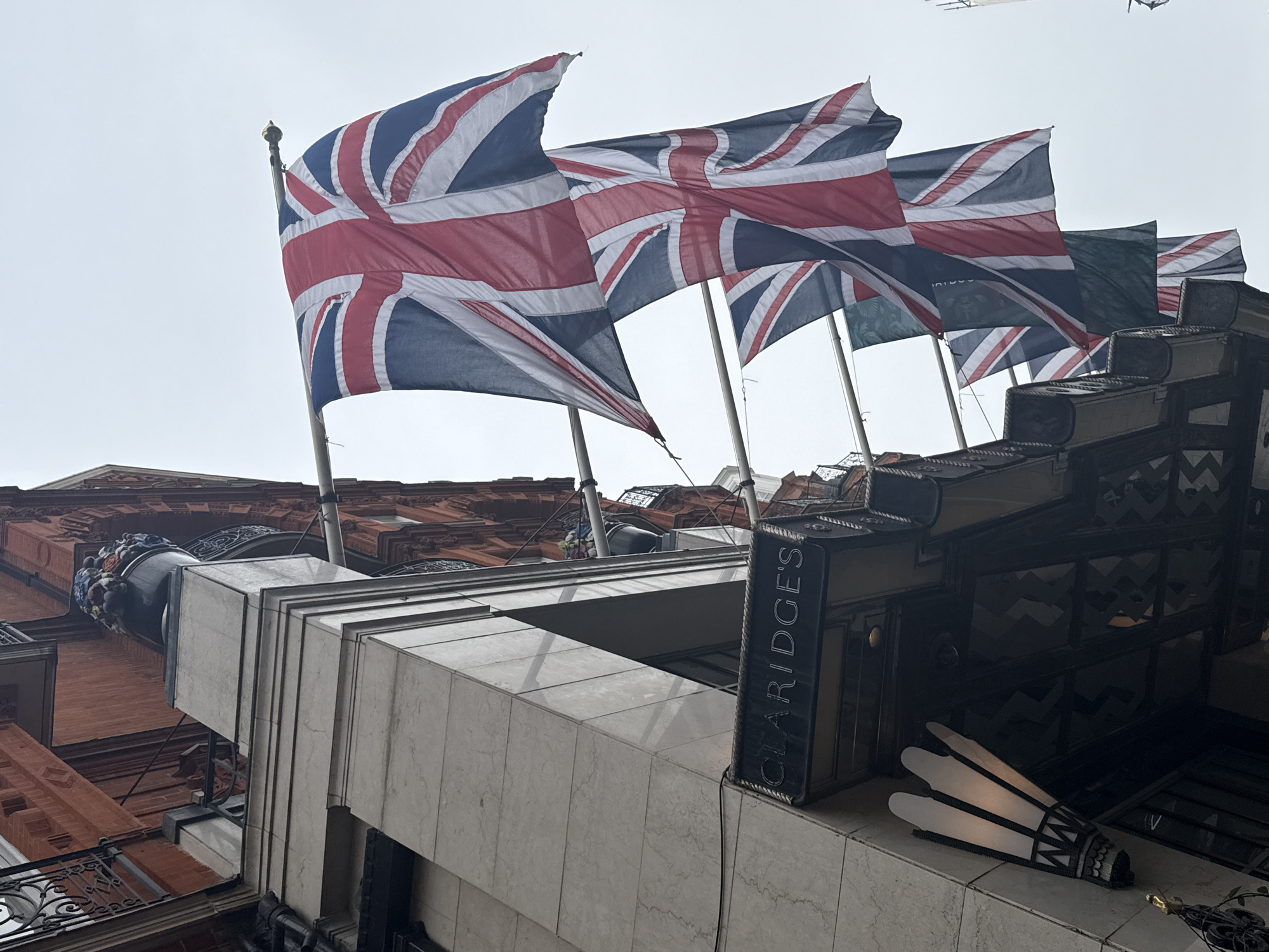 The image captures a low-angle view of Claridge's hotel in London, United Kingdom. Several Union Jack flags are flying from the roof, indicating a British theme or celebration. The flags are flapping in the wind, revealing the red, white, and blue of the British flag. Below the flags, the hotel's architectural details are visible, including the facade, the name "Claridge's" on a sign, and distinctive Art Deco-style lamps. The buildings are constructed of red bricks and white stone. The sky is overcast, giving the scene a muted tone. The perspective emphasizes the height and grandeur of the hotel.