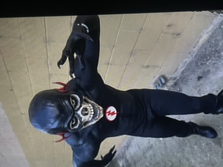 A single individual is depicted in an outdoor setting in Muchiya, Nigeria. The subject stands on a dark gray concrete surface, positioned in front of a light-colored cinder block wall. The individual wears a dark, full-body costume. This costume features a black full-head mask designed to resemble a skull, complete with white eye sockets, bared teeth, and red lightning-bolt shaped appendages at the temples. A prominent red circular emblem with a white lightning bolt is centered on the chest. Dark gloves with elongated, pointed fingertips adorn the hands, with white finger rings visible on the right hand. Dark boots are worn. The subject is captured leaning forward with both arms extended laterally, hands open with splayed fingers, suggesting a dynamic, poised stance.