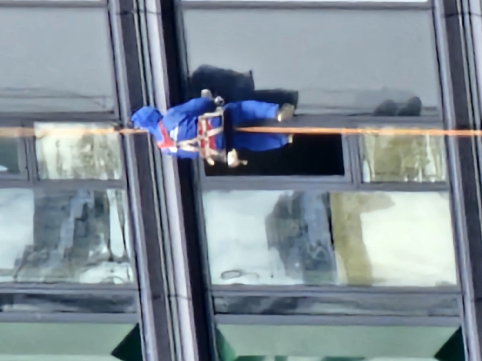 The image shows a window cleaner rappelling down the side of a modern building. The cleaner is wearing a blue jumpsuit and a safety harness. They are attached to a rope that is descending from above. The building appears to be made of glass and metal. The scene is captured from a side-on perspective, looking up at the cleaner as they work. The windows of the building reflect the sky and surrounding environment, suggesting an urban setting. The overall impression is one of a routine maintenance task on a commercial or residential high-rise.