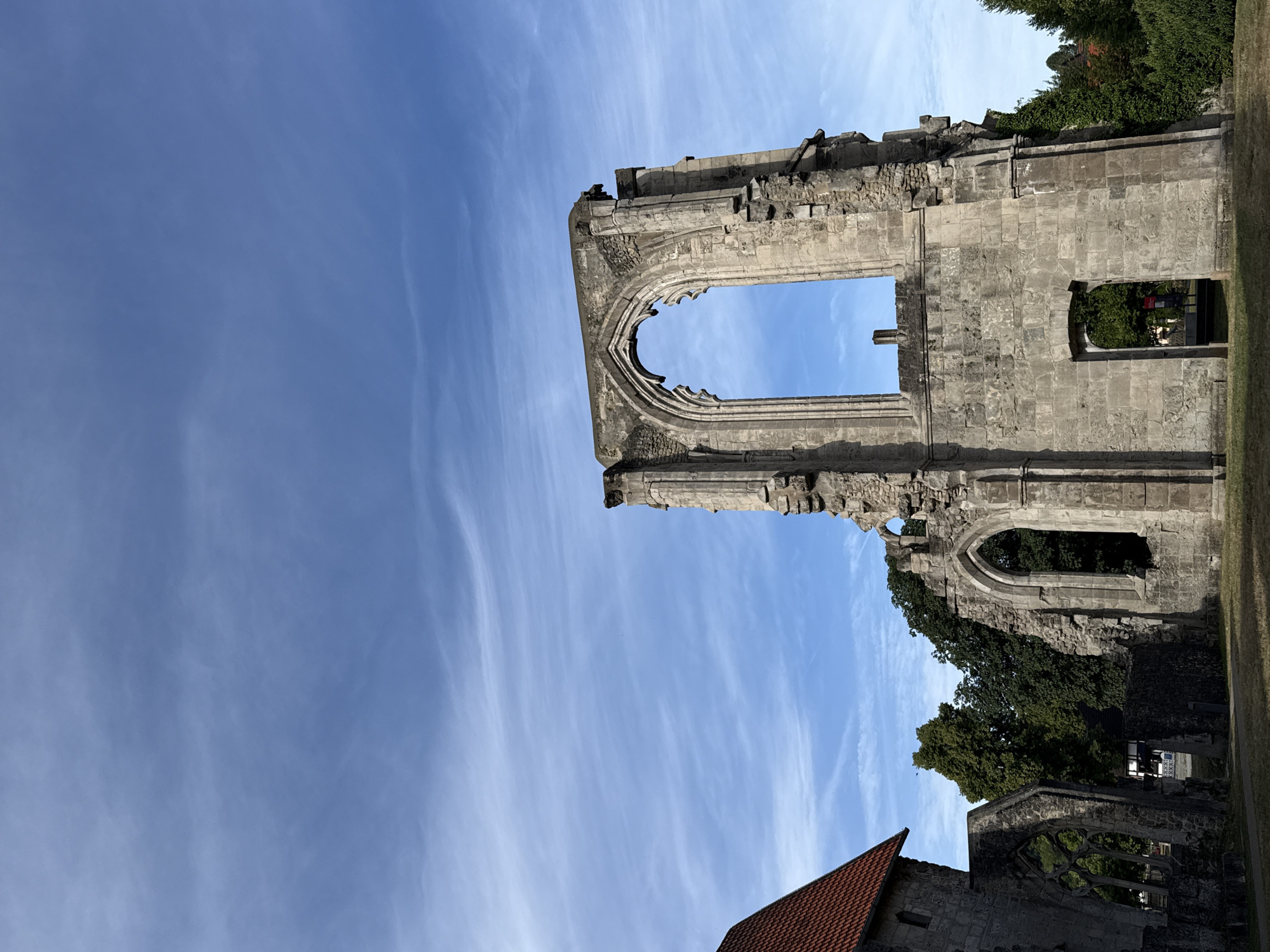 The image shows the ruins of a historic building, possibly a monastery, located in Walkenried, Germany. In the foreground, crumbling stone structures with arched windows dominate the scene. To the left, a partially intact building with a red-tiled roof is visible. In the background, lush green trees and a blue sky with wispy clouds create a serene atmosphere. The ruins appear weathered and aged, suggesting a long history. The overall impression is one of historical significance and natural beauty.