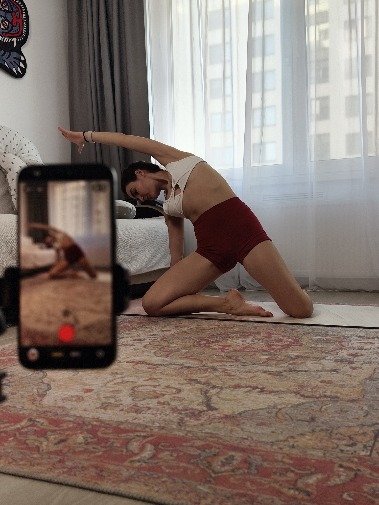 A woman is performing a yoga pose on a mat in a room, recording herself with a smartphone. The woman is wearing a white sports bra and maroon shorts. She is kneeling on the mat with her right knee on the ground and her left foot flat on the floor. Her right arm is raised overhead and bent at the elbow, while her left hand rests on her left thigh. The smartphone is mounted on a tripod and positioned in front of the woman, capturing her from the waist up. A red recording indicator is visible on the phone screen. Behind the woman is a sofa, a window with sheer white curtains, and buildings visible through the window. The scene is taking place indoors, likely in a home or apartment. The room is carpeted with a patterned rug. Given the architectural style of the buildings seen through the window, and based on the general layout of the room which suggests it could be a domestic interior, it is likely that this is in the city of Tyumen, Russia. The room is sparsely furnished and lit with natural light coming through the window.