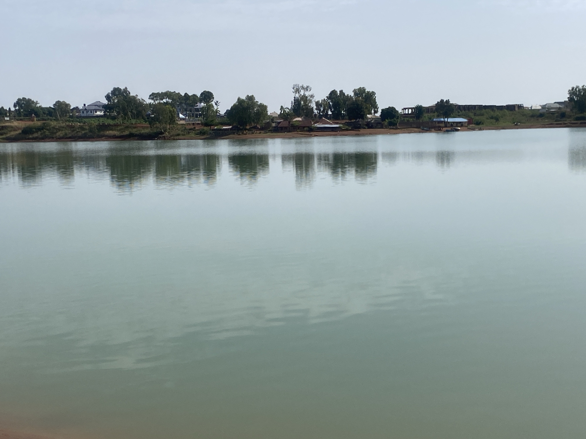 The image depicts a calm body of water, possibly a lake or wide river, reflecting the landscape on the opposite shore. The water is a light, murky green color, with subtle ripples and reflections. The far shore features a line of trees, interspersed with buildings, some of which appear to be houses with light-colored roofs. Further back, there's a larger structure under construction. The sky is overcast and a pale, washed-out blue. The location is Unknown, Unknown.
