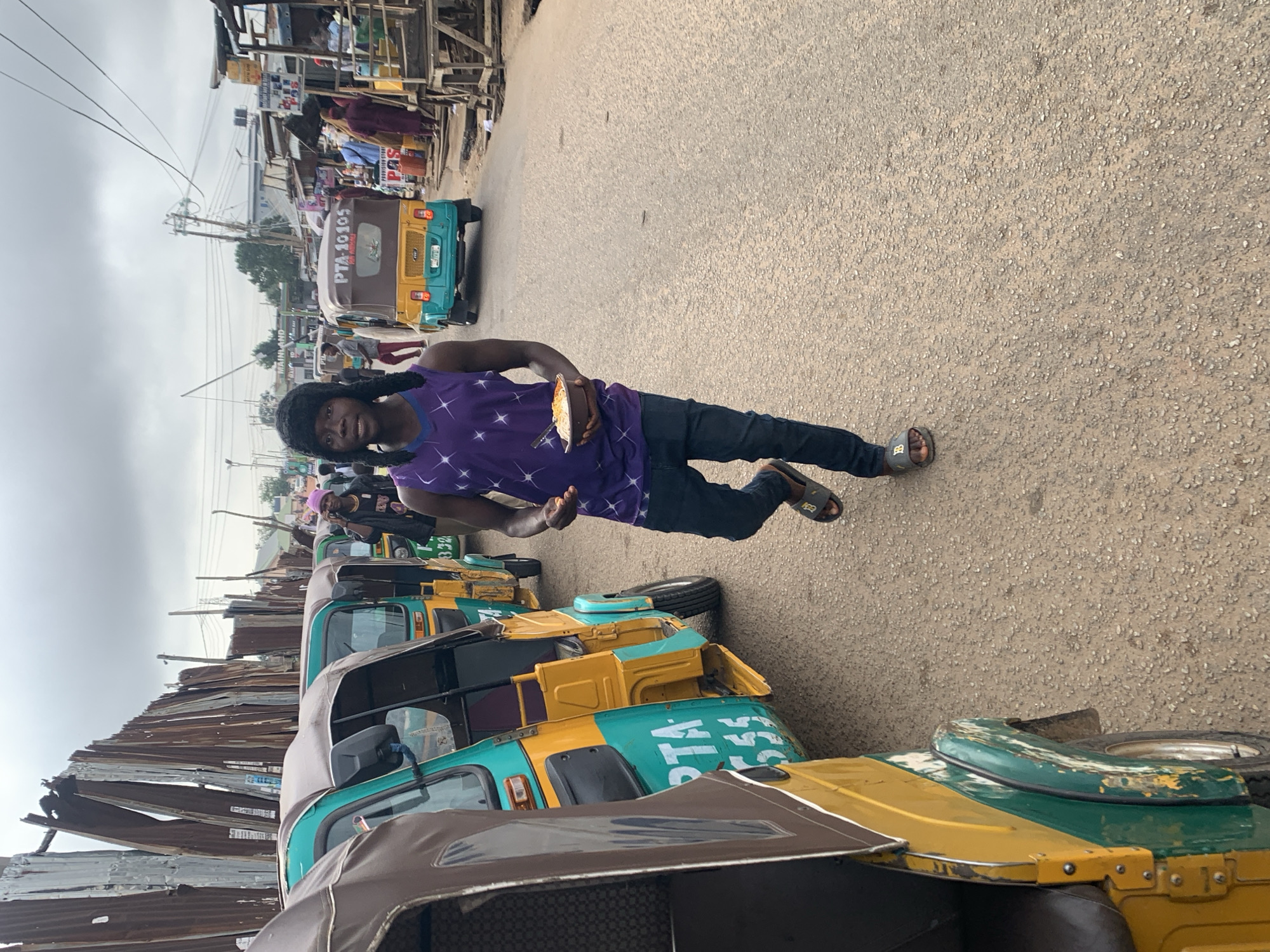 A young Black male, wearing a purple tank top with white star-like patterns and dark blue jeans, walks down a dusty, paved road in Jos, Nigeria, holding a bowl of food in his right hand. He is facing the camera and has a dark, fuzzy hat on his head, with braided extensions hanging down. He is wearing dark sandals. To his left, a line of yellow and teal auto-rickshaws, also known as Keke Napeps, are parked along the side of the road. The rickshaws are a common mode of transportation in Nigeria. In the background, there are various shops and buildings made of corrugated metal and wood, indicating a bustling market or commercial area. The sky is overcast with gray clouds. The overall scene depicts a typical street view in a Nigerian city.