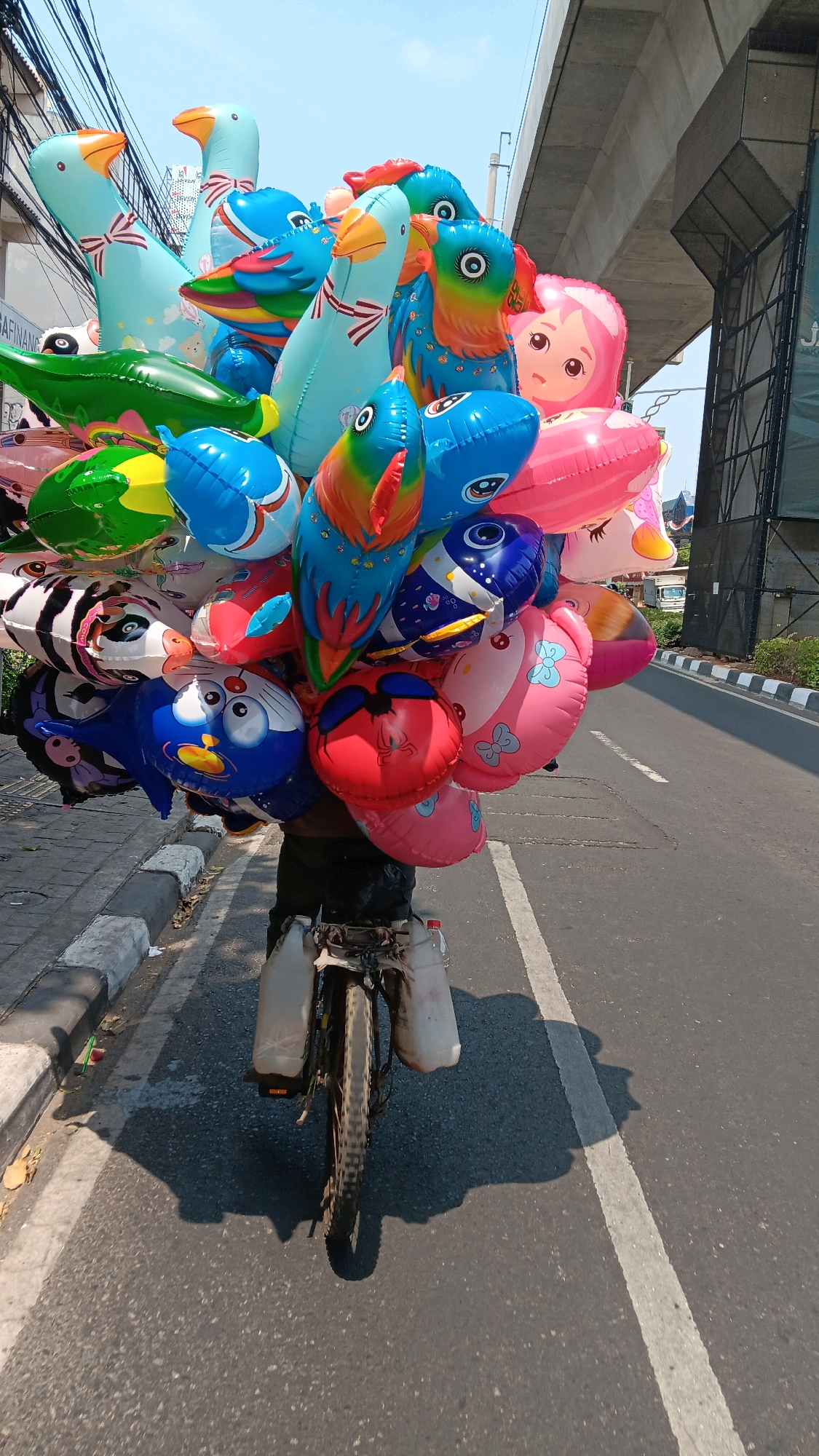 The image shows a person riding a bicycle laden with a large bunch of colorful balloons on a city street. The balloons are in various shapes and characters, including ducks, birds, and possibly cartoon characters. The bicycle is equipped with two large plastic containers attached to the rear rack, which appear to be filled with water. The street has a sidewalk with a curb and is paved with asphalt. In the background, there is a concrete overpass or bridge structure. The lighting suggests it is daytime with bright sunlight, casting shadows on the road. The environment appears to be urban, potentially in South Jakarta, Indonesia, given the context provided. The overall scene depicts a street vendor selling balloons from a bicycle.