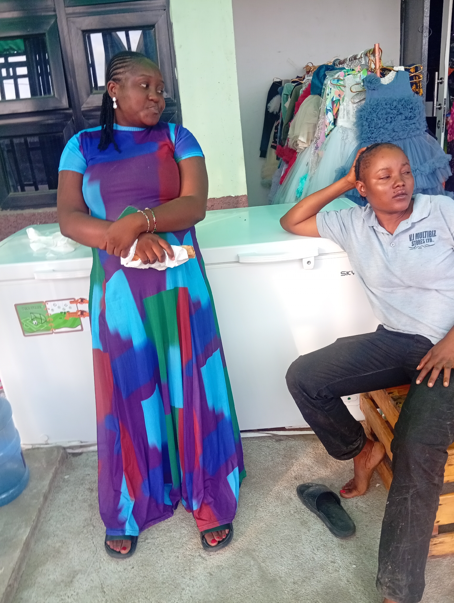 Two women are standing and sitting in front of a chest freezer and a rack of dresses. The woman on the left is standing and wearing a colorful, long dress with blue, purple, red, and green abstract patterns. She has her arms crossed and is holding a white napkin with something inside. She has braided hair and is wearing earrings and bracelets. The woman on the right is sitting on a wooden stool, leaning against the chest freezer. She is wearing a grey polo shirt with a logo and black pants. Her head is tilted back and her eyes are closed. A black sandal is on the floor near her foot. The chest freezer has stickers on the front, one of which appears to be a soccer-themed graphic. Behind the women, there is a rack with several dresses hanging on it. The scene appears to be outdoors, possibly in a market or shop, given the display of clothing and the presence of a water dispenser in the background. The lighting suggests it is daytime. The woman on the right seems relaxed, while the woman on the left has a more engaged expression. Visible text on the grey shirt reads "V.I. MULTIBIZ STORES LTD.". The location is identified as Howayi, Nigeria.