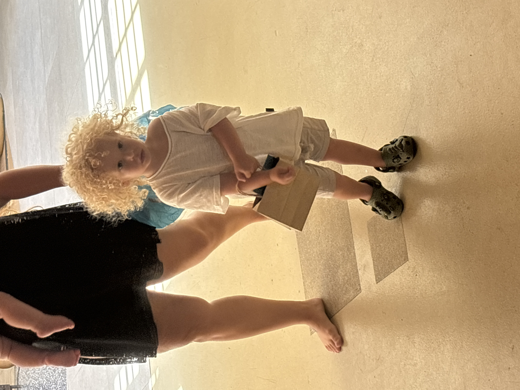 A blonde curly-haired child wearing a white t-shirt, gray shorts, and dark-colored Crocs is standing on a tiled floor holding a rectangular object. An adult wearing black shorts is standing near the child. The location is presumed to be indoors. Based on the request, the location is Baan Bo Phut, Thailand. The child is looking directly at the camera. Natural light is visible from an unseen window to the left of the child. The floor tiles are light brown. The lighting is bright.