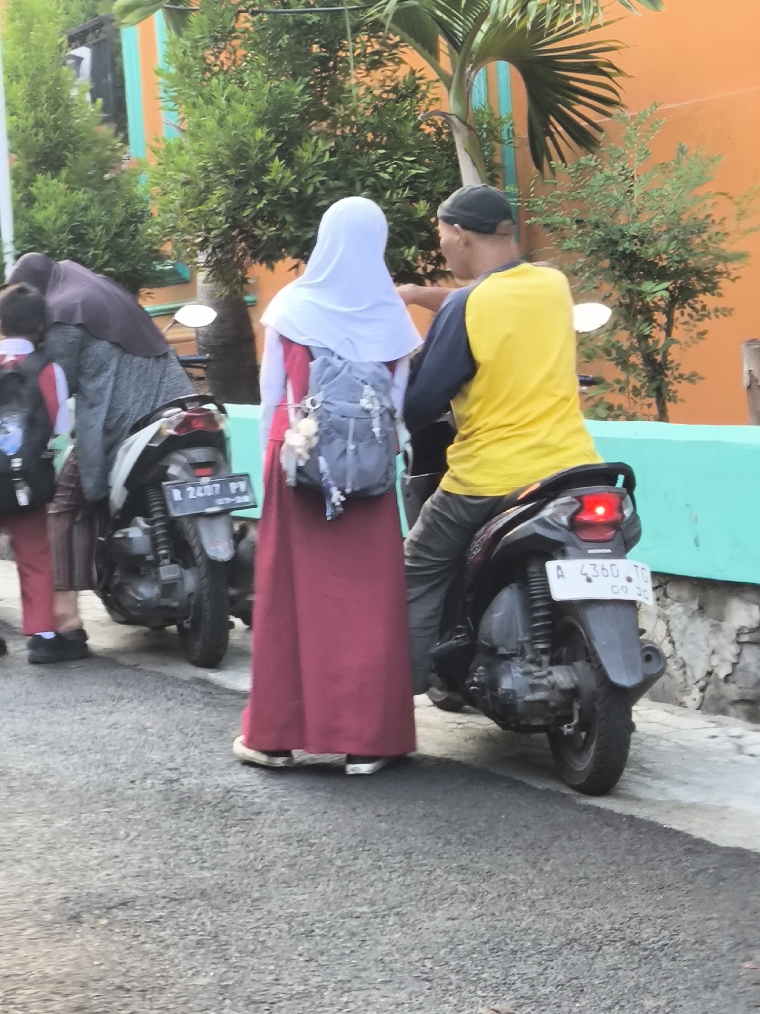 In the city of Cilegon, Indonesia, two individuals are interacting near parked motorcycles. On the right, a person wearing a yellow shirt with dark sleeves and a dark cap is seated on a black motorcycle with a red brake light illuminated. Their legs are visible, clad in grey trousers. To the left, a person wearing a white hijab and a long, maroon dress is standing next to a grey backpack with decorative charms. Partially visible to the left of the person in the maroon dress is another individual wearing a dark hijab and a grey outfit, standing beside a motorcycle. The ground is paved with asphalt, and a turquoise wall is present in the background, partially obscured by green foliage and an orange building. A motorcycle license plate reads "R 2487 PV". Another license plate on the motorcycle to the right reads "A 4360 TG 09 20".