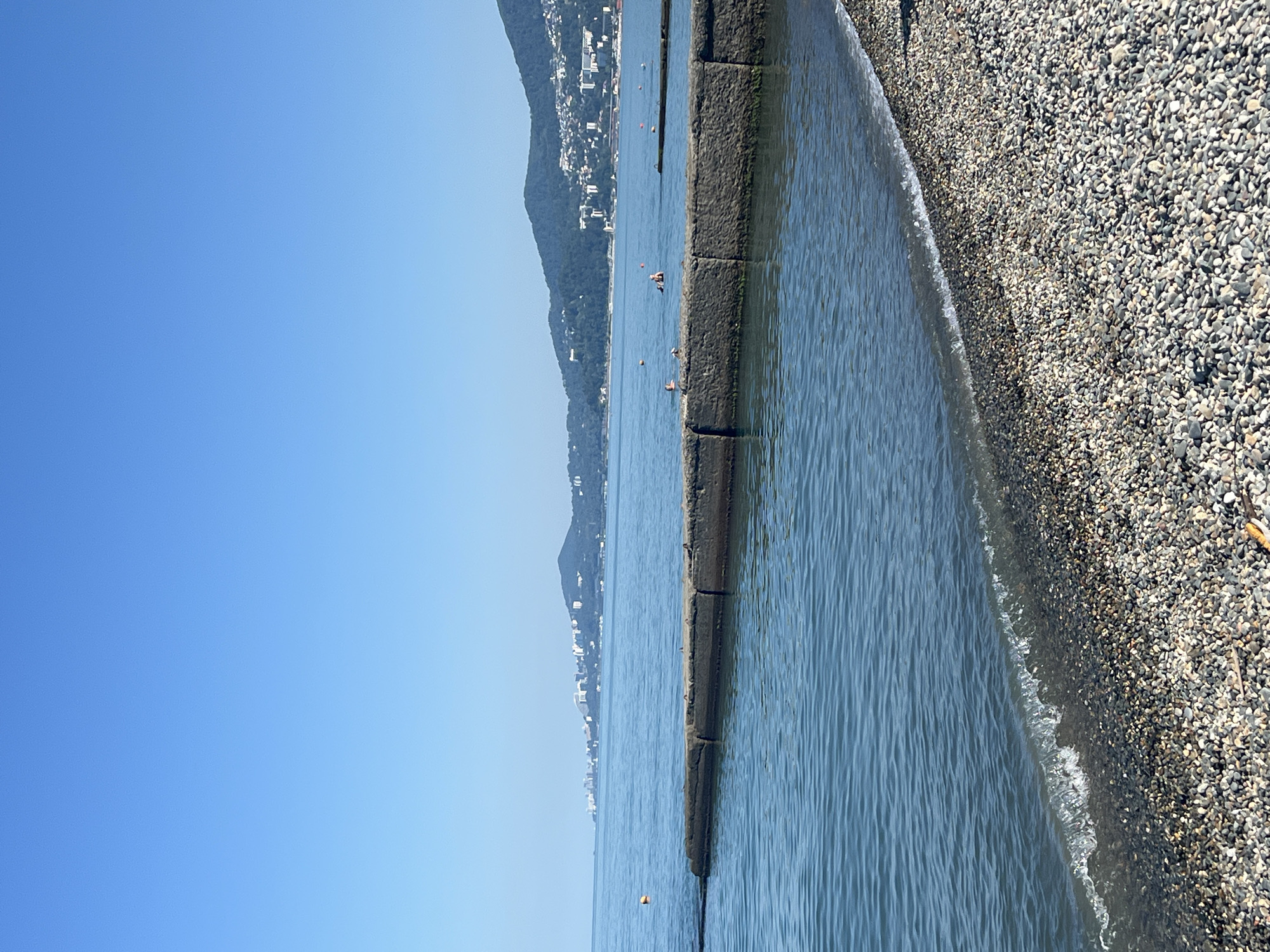 This image captures a serene coastal scene in Sochi, Russia, likely during the daytime under a clear blue sky. In the foreground, a pebbled beach slopes gently towards the calm, blue waters of the Black Sea. A low, concrete breakwater, constructed from rectangular blocks, extends into the water from the left, creating a sheltered area. Beyond the breakwater, the sea stretches towards a distant coastline characterized by rolling, tree-covered hills dotted with white buildings. The overall impression is one of a tranquil summer day at a seaside resort.