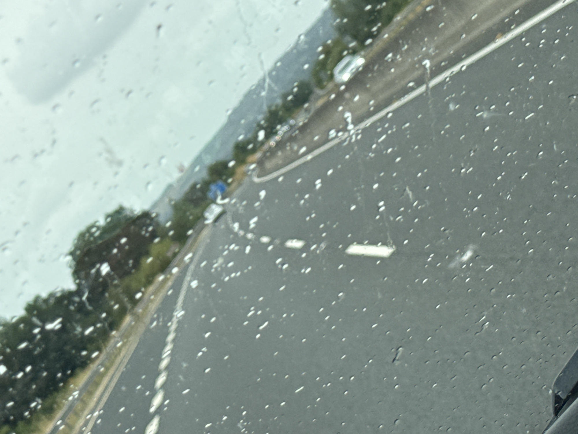 The image depicts a view from inside a vehicle, looking through a wet windshield at a highway. The windshield is covered in numerous raindrops, which distort the view of the road and surrounding landscape. The highway appears to be asphalt, with white lane markings visible. There are a few vehicles on the road, but they are blurred and indistinct due to the raindrops and motion. The landscape beyond the highway consists of trees and a hillside, all softened by the rain and the wet glass. The sky is overcast and grey. Based on the provided information, the location is identified as a highway in the vicinity of Craan, Ireland.