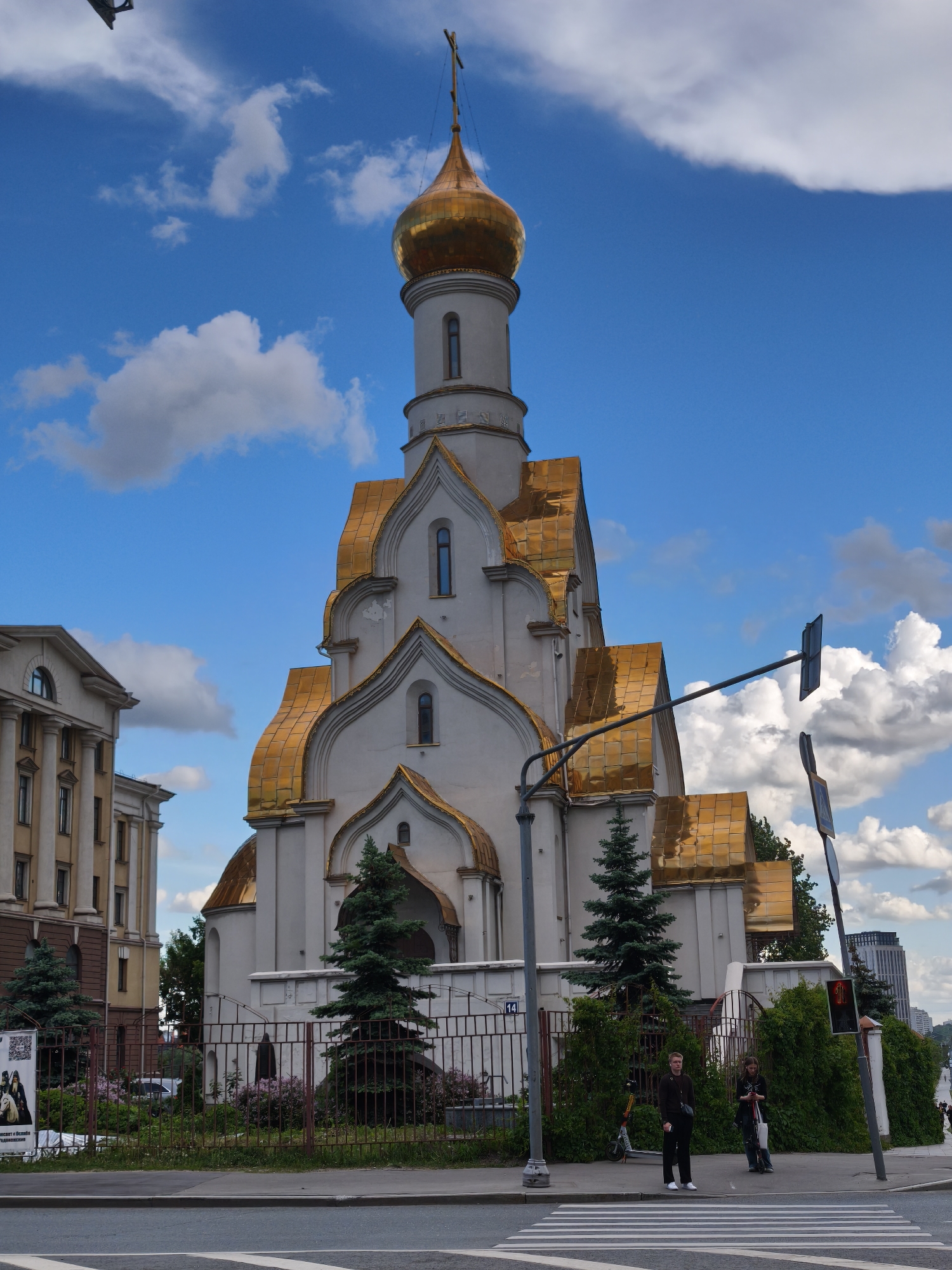 The image captures the Church of All Saints in Moscow, Russia, set against a blue sky dotted with cumulus clouds. The church's architecture features a central tower topped with a golden dome and a cross. The roof is also covered in golden tiles. The main structure is light-colored, likely white or light gray.
A low metal fence surrounds the church grounds, and greenery is visible, including trees and bushes. In front of the church, a crosswalk stretches across the street.
Two people are standing near the crosswalk on the sidewalk. One person is wearing dark clothing, and the other person is holding a scooter. Traffic lights and street signs are positioned nearby.
To the left of the church, part of another building is visible, constructed with pale stone and multiple windows. The overall setting suggests an urban environment.
