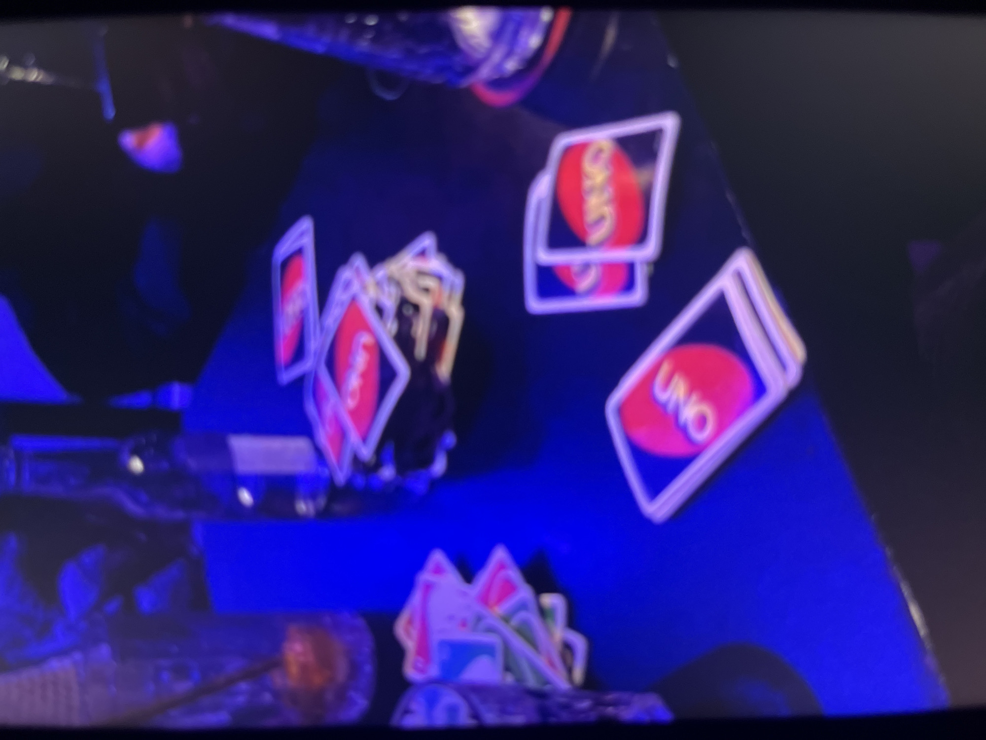 The image depicts a table with several UNO cards scattered across it, illuminated by blue lighting. A glass with a straw and an orange-colored liquid is visible on the left side of the table, along with a bottle that appears to be a beverage. A stack of UNO cards is in the center of the table, and two individual UNO cards are in the foreground. The scene suggests a social gathering or game night. The location of Bukuru, Nigeria, is not visually discernible from the provided image.