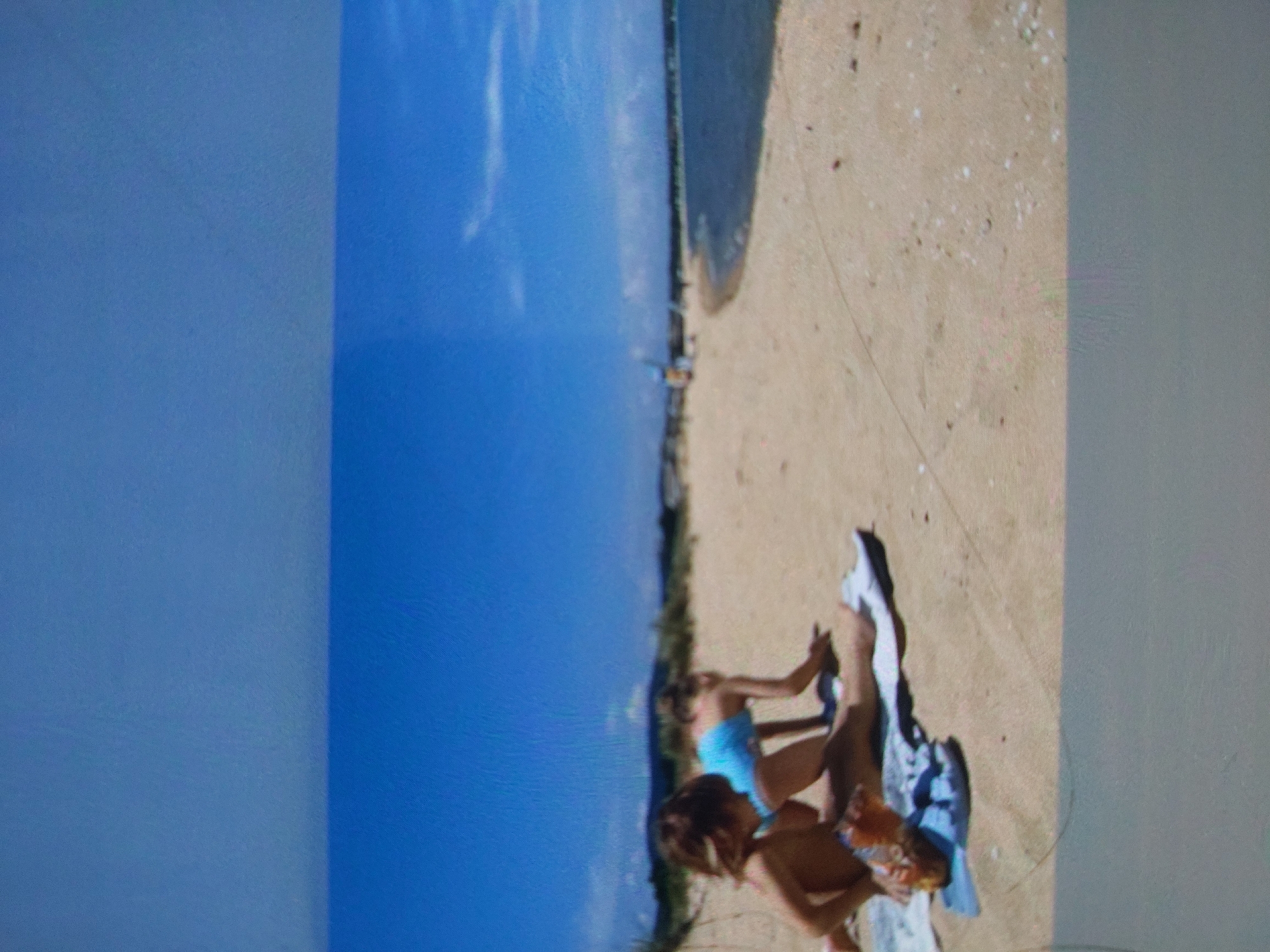 The image shows a beach scene in Tsarevo, Bulgaria. The beach is sandy, with some vegetation along the edge. Two people, possibly children, are sitting on the sand. One is wearing a blue swimsuit. There is a towel or piece of fabric next to them. The sky is blue and clear.