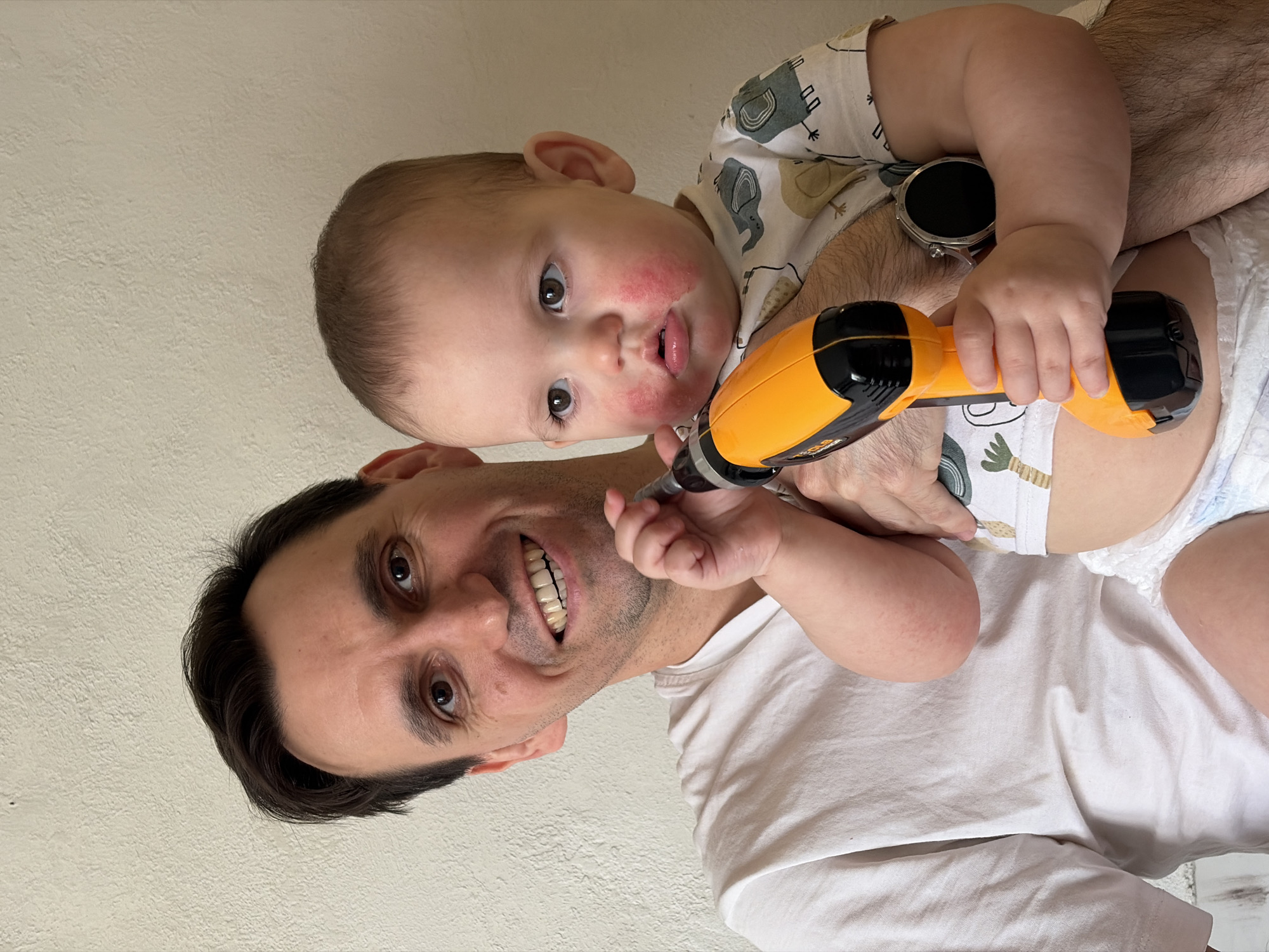 A man with dark hair and a white t-shirt is holding a baby in his arms. The baby has reddish patches on its face and is wearing a printed white shirt and a diaper. The baby is holding a black and yellow power drill. The man is smiling. The background is a plain wall. The image does not give indication that it was taken in Chaikovsky, Russia.