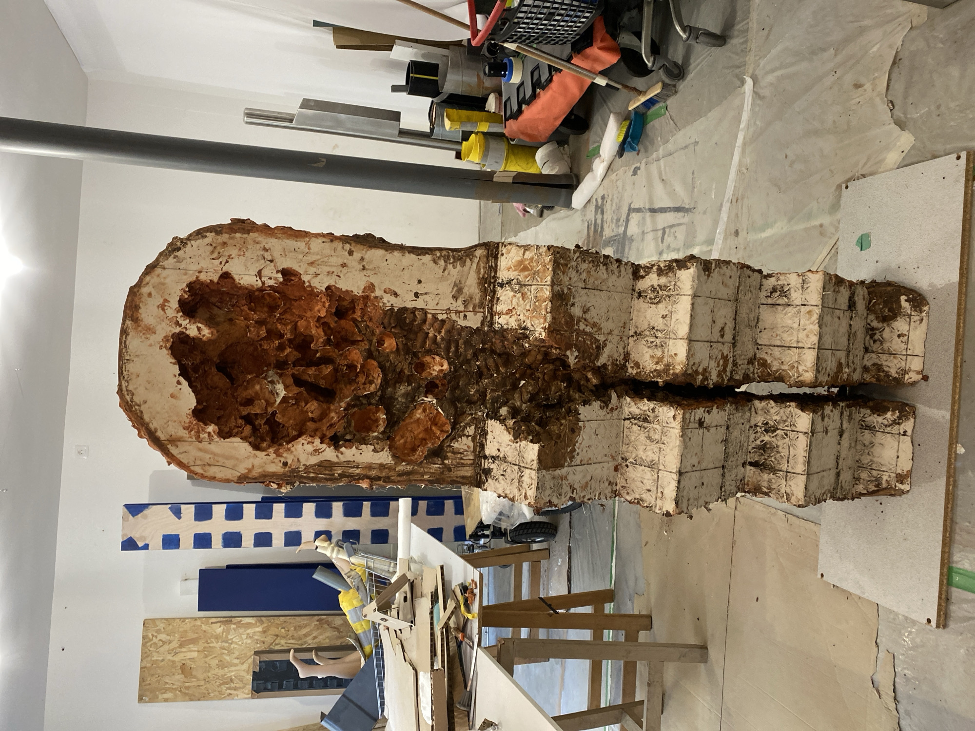The image shows a large, rough-hewn sculpture in a cluttered indoor space. The sculpture is vertical, resembling a hollowed-out, elongated form with a reddish-brown, cavernous interior and a textured, off-white exterior with blocky segments. It is split lengthwise down the middle. The surrounding environment appears to be a workshop or storage area with various tools, materials, and discarded objects. There is a wooden structure nearby, possibly a work table or support. The space has a utilitarian feel, with white walls and a light-colored floor covered with a protective sheet. There is no indication that the location is in Paris, France. No people are visible in the image.