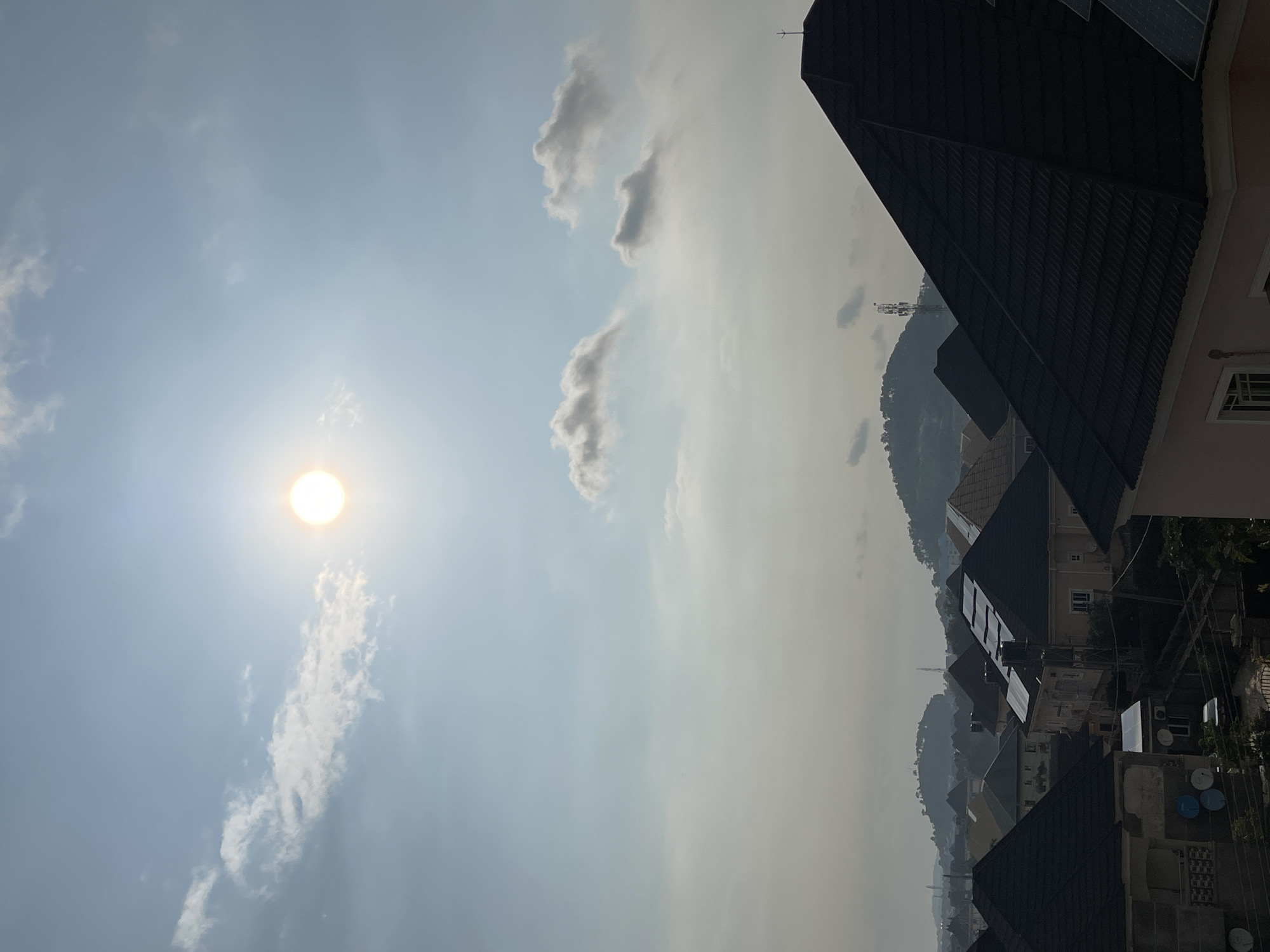 This is a landscape photograph taken from a high vantage point, overlooking a residential area in Abuja, Nigeria. The sky is bright, with the sun prominently positioned in the upper left quadrant, casting a soft glow and appearing slightly hazy due to atmospheric conditions. Scattered clouds drift across the sky, some appearing white and fluffy, others with a slightly gray hue.
In the foreground, the roofs of several houses are visible. These are primarily dark-colored, tiled roofs, with some featuring solar panels. The architecture suggests a modern residential development. Beyond the immediate houses, the landscape recedes into a series of rolling hills, also covered in vegetation and appearing somewhat distant and muted due to the haze. A cell tower is visible on one of the hills, indicating infrastructure within the broader urban or semi-urban environment.
The scene is devoid of people or any visible human activity. The overall impression is one of a quiet, residential neighborhood under a bright sky, possibly during the morning or late afternoon given the sun's position. The image captures a typical view of a developed area in Abuja, characterized by its distinct topography and modern housing.