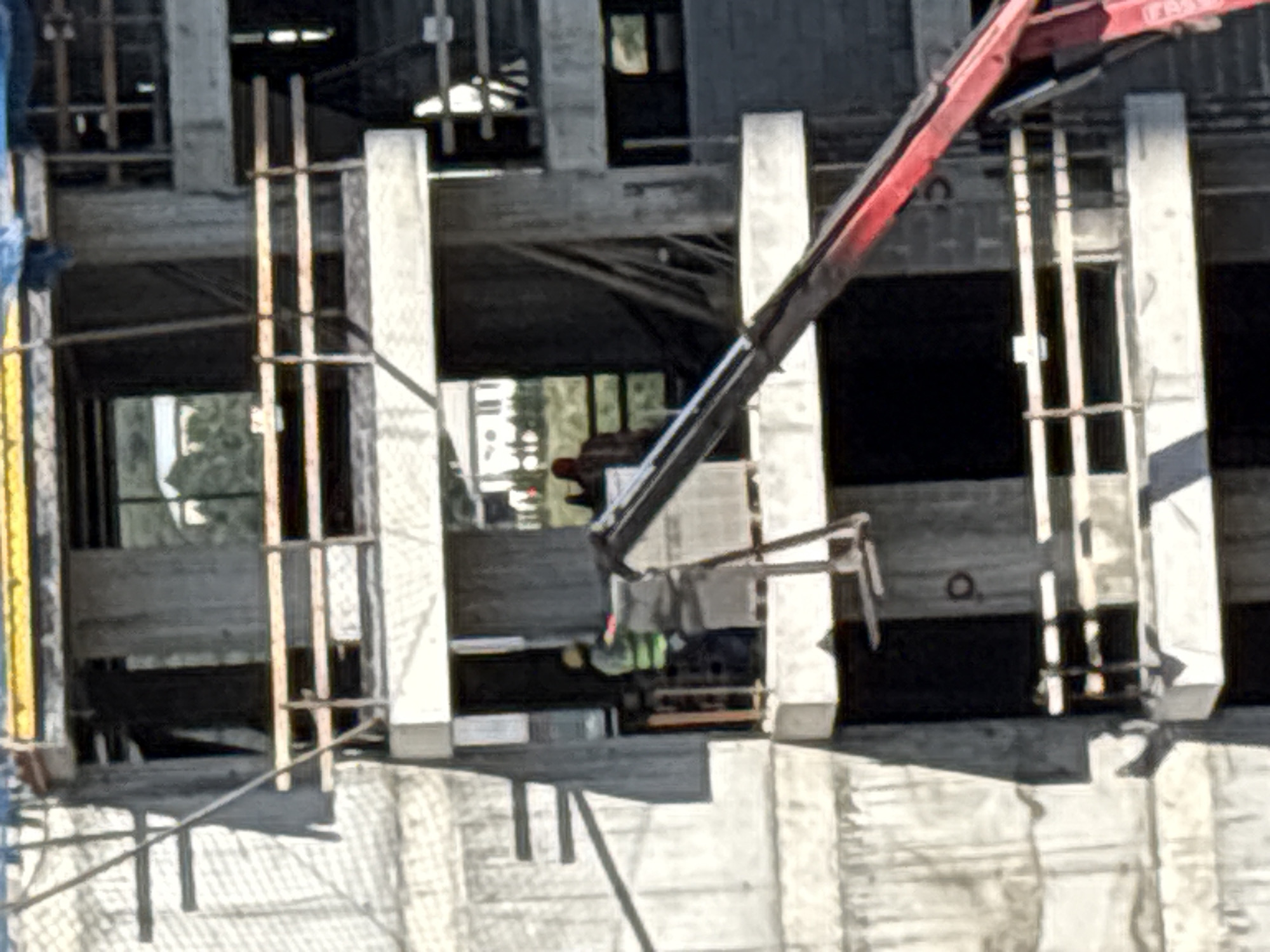 The image captures a construction site in Tbilisi, Georgia, focusing on the exterior of a multi-story building under development. The building's structure is primarily concrete, with visible scaffolding and safety railings made of wood and metal. On one of the building's levels, two construction workers are present. One worker, wearing a bright green safety vest and what appears to be a yellow hard hat, is positioned near an opening. Another worker, dressed in a brown garment, is also visible within the building's structure. A red crane arm extends into the frame from the right side, suggesting ongoing lifting or placement of materials. The surrounding environment shows glimpses of other buildings and greenery through the open sections of the construction site. The overall scene depicts an active construction environment in an urban setting.