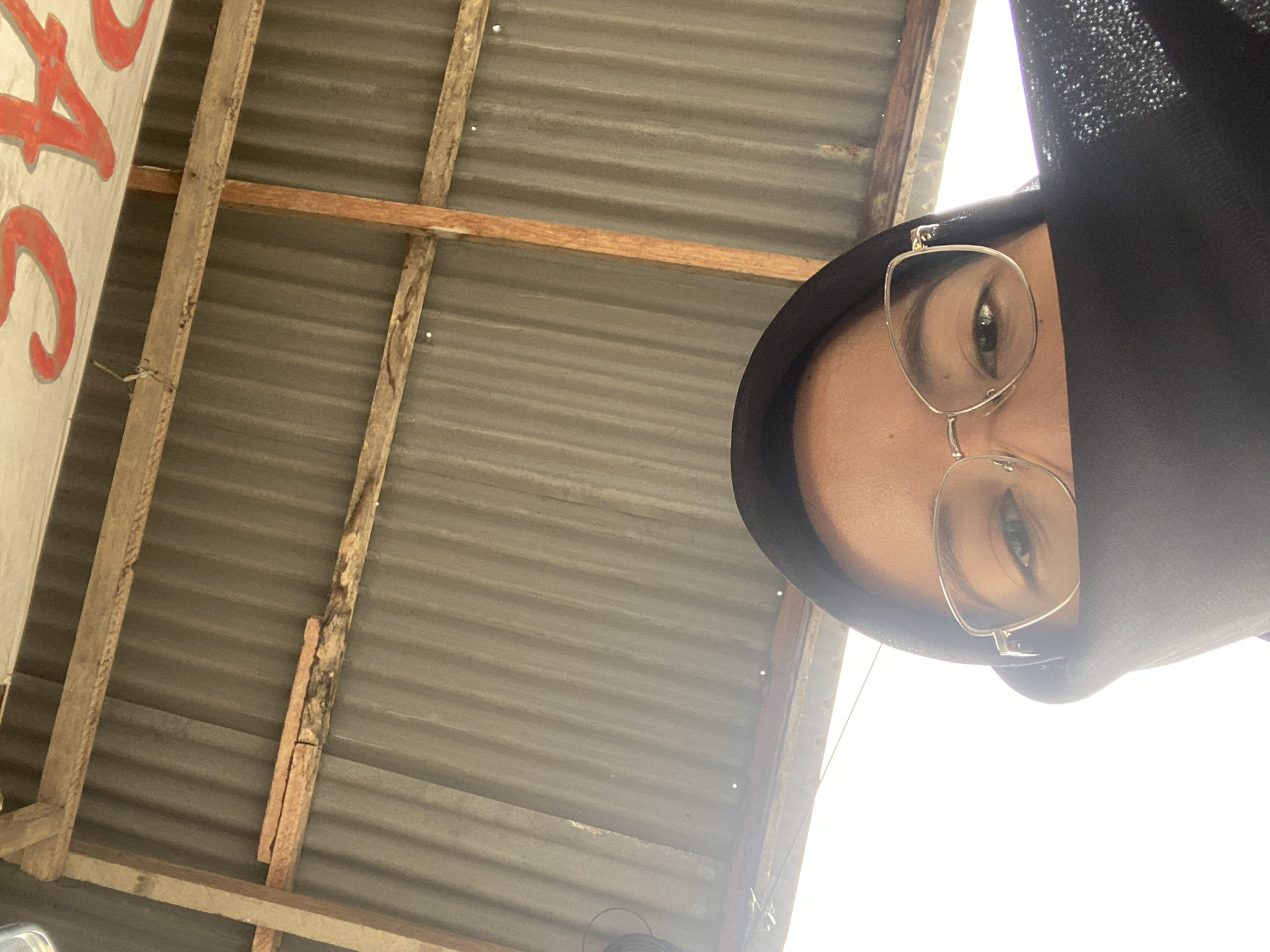 A close-up, overhead, indoor, selfie-style shot shows a person wearing a black hijab and glasses. The person's face is partially obscured by the hijab, revealing only their eyes, eyebrows, and the bridge of their nose. They are wearing gold-rimmed, cat-eye glasses. The background consists of a corrugated metal roof supported by wooden beams. Red lettering is visible on a white surface above the roof. The scene appears to be within a building or outdoor shelter in Air Molek, Indonesia. The lighting suggests an overcast day or diffused natural light.